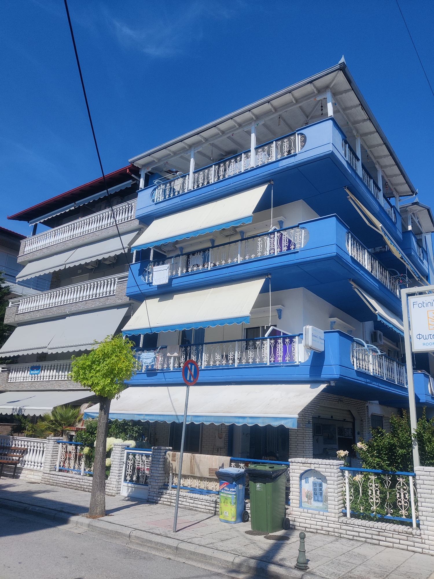 The image shows a building in the city of Katerini Municipality, Greece, on a sunny day. The building is painted bright blue and white and has multiple balconies with white railings. White awnings are extended over some of the balconies. A tree with green leaves stands in front of the building. A sidewalk and a street are visible in the foreground, with two trash bins and a signpost on the sidewalk. The sky is clear and blue.