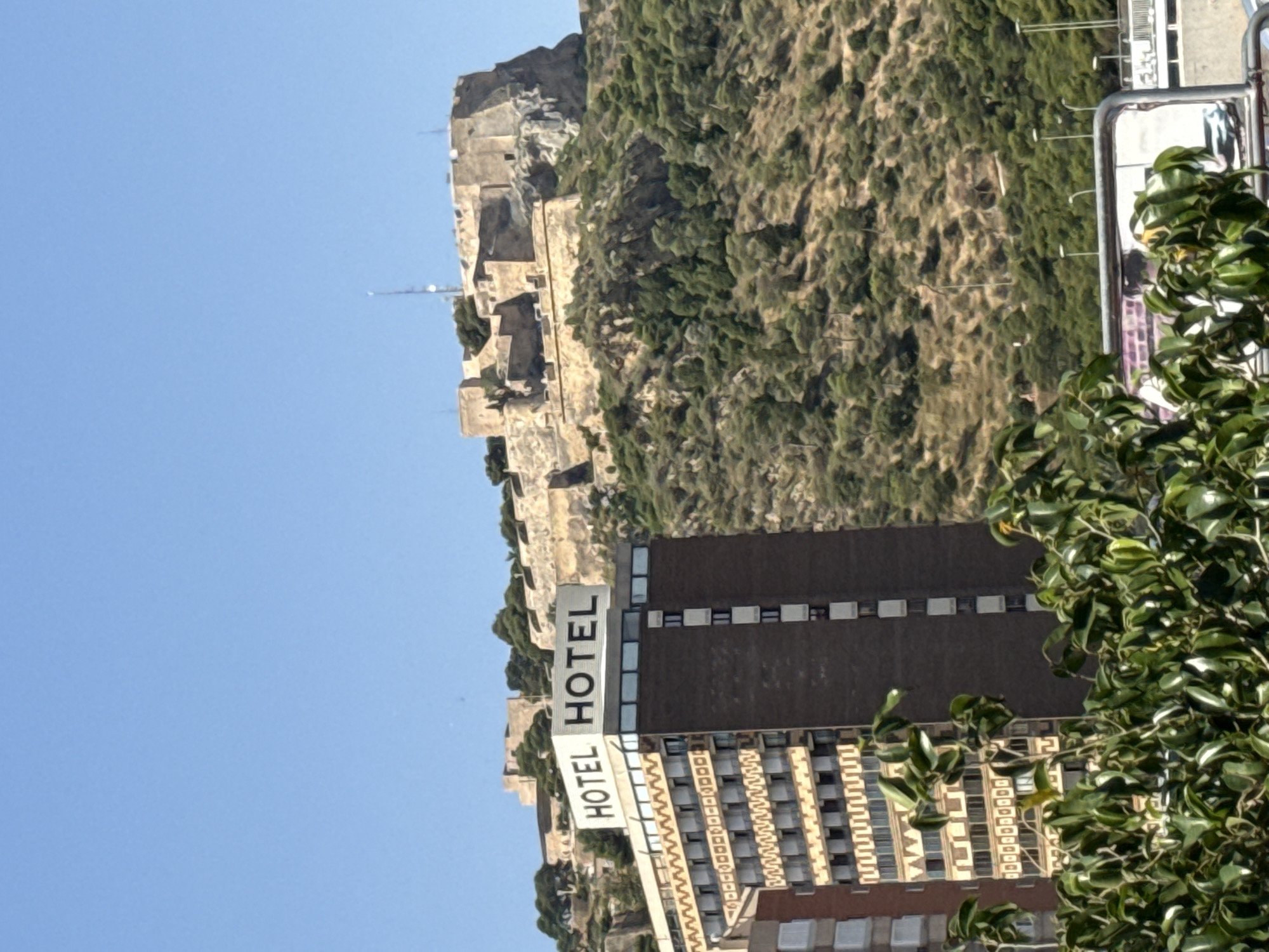 A sunny, clear day illuminates a landscape featuring a large, verdant mountain or hill in the background. The mountain is densely covered with green trees and shrubs, and ancient stone structures, resembling a fortress or castle ruins, are visible on its summit and integrated into its slopes. A slender antenna or communication mast rises faintly from the mountain's peak against the blue sky.
In the midground, in front of the mountain, stand modern buildings. To the right, a tall, dark-colored building with multiple windows, likely a hotel, is prominent. To its left, another large building, also appearing to be a hotel, features a distinctive, patterned facade. A prominent white sign on the side of this building clearly displays the word "HOTEL" twice in large, black capital letters.
The immediate foreground on the right is occupied by lush green foliage from a tree or bush, partially obscuring the modern buildings. A silver-colored metal railing or structure is partially visible in the upper right corner, behind the foreground foliage. The sky is a cloudless, bright blue. No people are visible in the static scene.