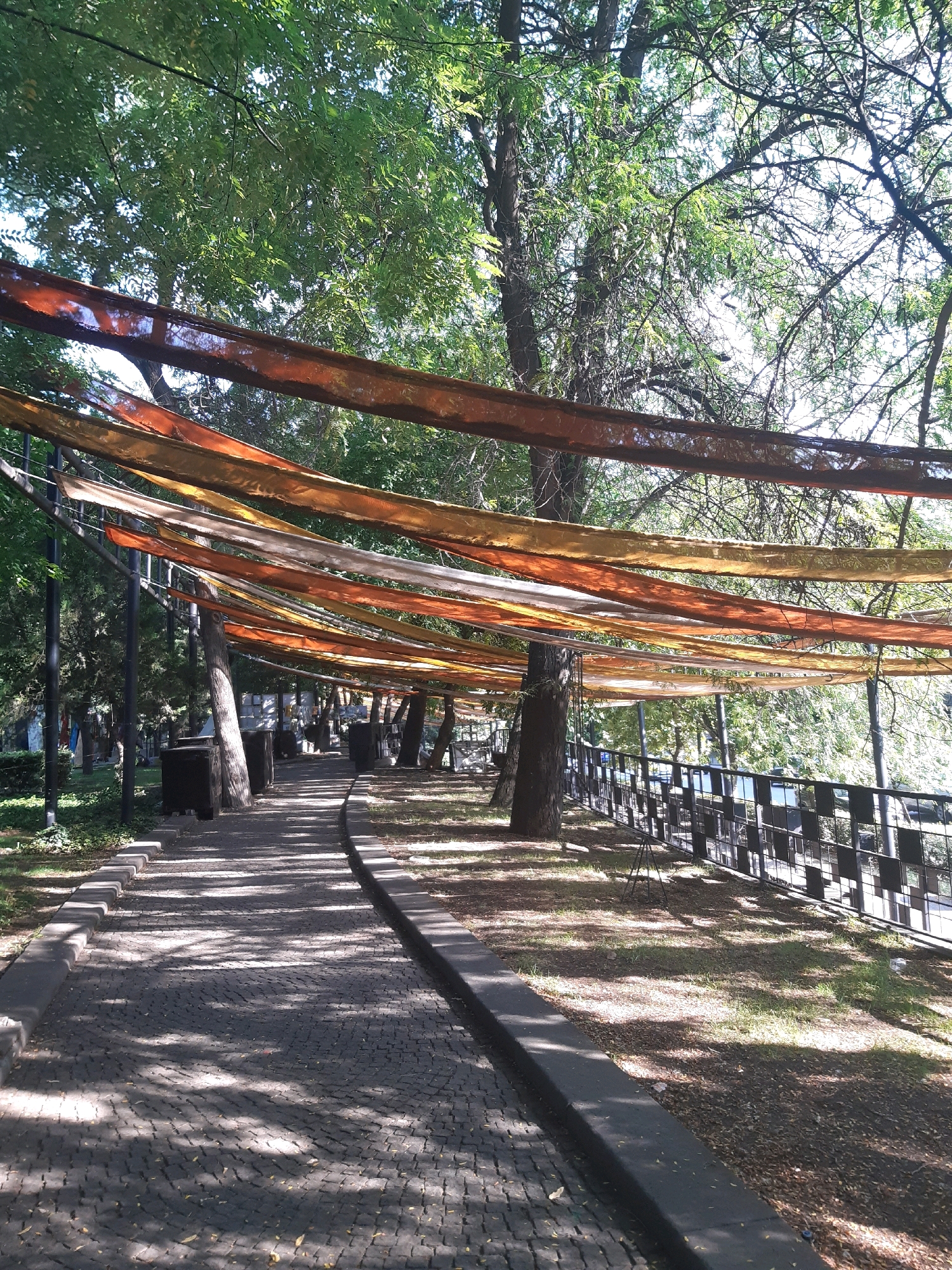 The image shows a park pathway in Tbilisi, Georgia, with colorful fabric strips draped overhead, creating a canopy. The pathway is paved with cobblestones and is lined by trees and a metal fence. Sunlight filters through the leaves of the trees, casting dappled shadows on the ground. The scene appears peaceful and inviting, suggesting a pleasant day in the park.