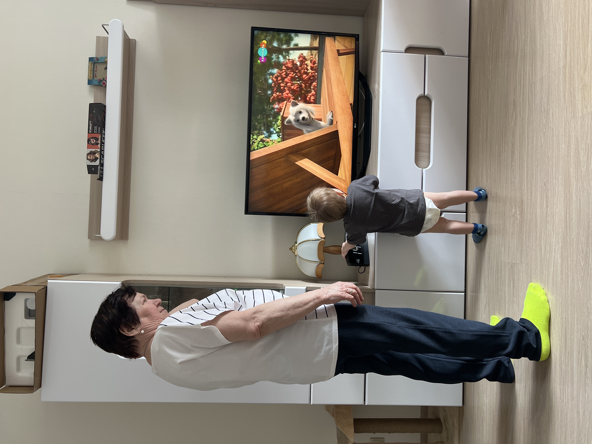 The image captures an indoor scene, likely a living room, featuring an older woman and a young child. The room has light-colored walls and light wood flooring, illuminated by natural or overhead indoor lighting, suggesting daytime.
An older woman, with short dark hair and pearl earrings, stands on the left side of the frame. She wears a white t-shirt with thin black stripes, dark blue trousers, and bright yellow socks. Her left arm is bent at the elbow, and she wears a ring on her left ring finger. She is looking towards the right, away from the camera.
A young child, appearing to be a toddler, stands closer to the center-right. The child has light-colored hair and wears a dark grey top, a diaper, and blue shoes. They are focused on a black flat-screen television and appear to be holding a small black remote control or device, interacting with a black object on the white TV stand directly below the screen.
The television displays an animated scene featuring a white dog looking over a wooden railing, with green foliage and red berries in the background. In the top left corner of the screen, the Russian text "Карусель" (Carousel, a children's TV channel) is visible.
Below the television is a white TV stand or cabinet. To the left of the child, on the TV stand, is a decorative table lamp with a pleated beige shade and a wooden base. Above the lamp and to the left of the TV, a light wood floating shelf is mounted on the wall. On this shelf, there is a small book or picture frame with a blue cover and a black box labeled "SCARLETT" featuring images of three women. On the far left of the image, an open cardboard box containing white packaging material rests on top of another white cabinet. The overall setting is domestic and quiet.