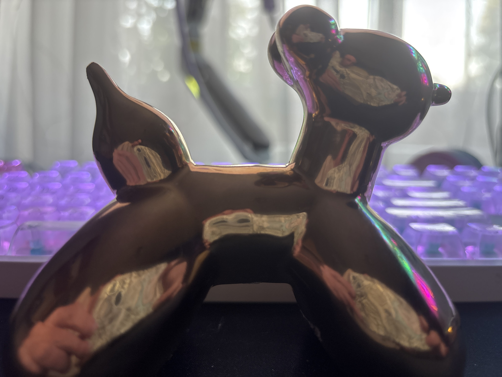 A close-up, eye-level shot features a metallic, balloon-dog figurine on a surface. The figurine is dark and highly reflective, showing distortions of its surroundings. Behind the figurine, a keyboard with illuminated purple keys is visible, suggesting an indoor setting. The background is out of focus with soft, blurry lights, hinting at a window or a brightly lit area. The overall lighting is dim with colorful reflections on the figurine.