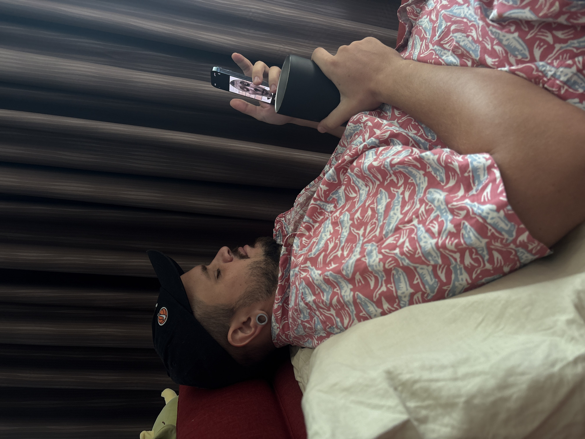A man lies on a white pillow on a red sofa. He is wearing a black baseball cap with an orange basketball emblem and a red and white patterned shirt with sharks. He is holding a black cylindrical object with a smartphone attached to it, possibly watching something on the screen. The background is a brown striped wall. The image suggests an indoor setting, possibly a home or hotel in Choeng Thale, Thailand. No specific indicators or landmarks are visible to confirm the exact location within the city.