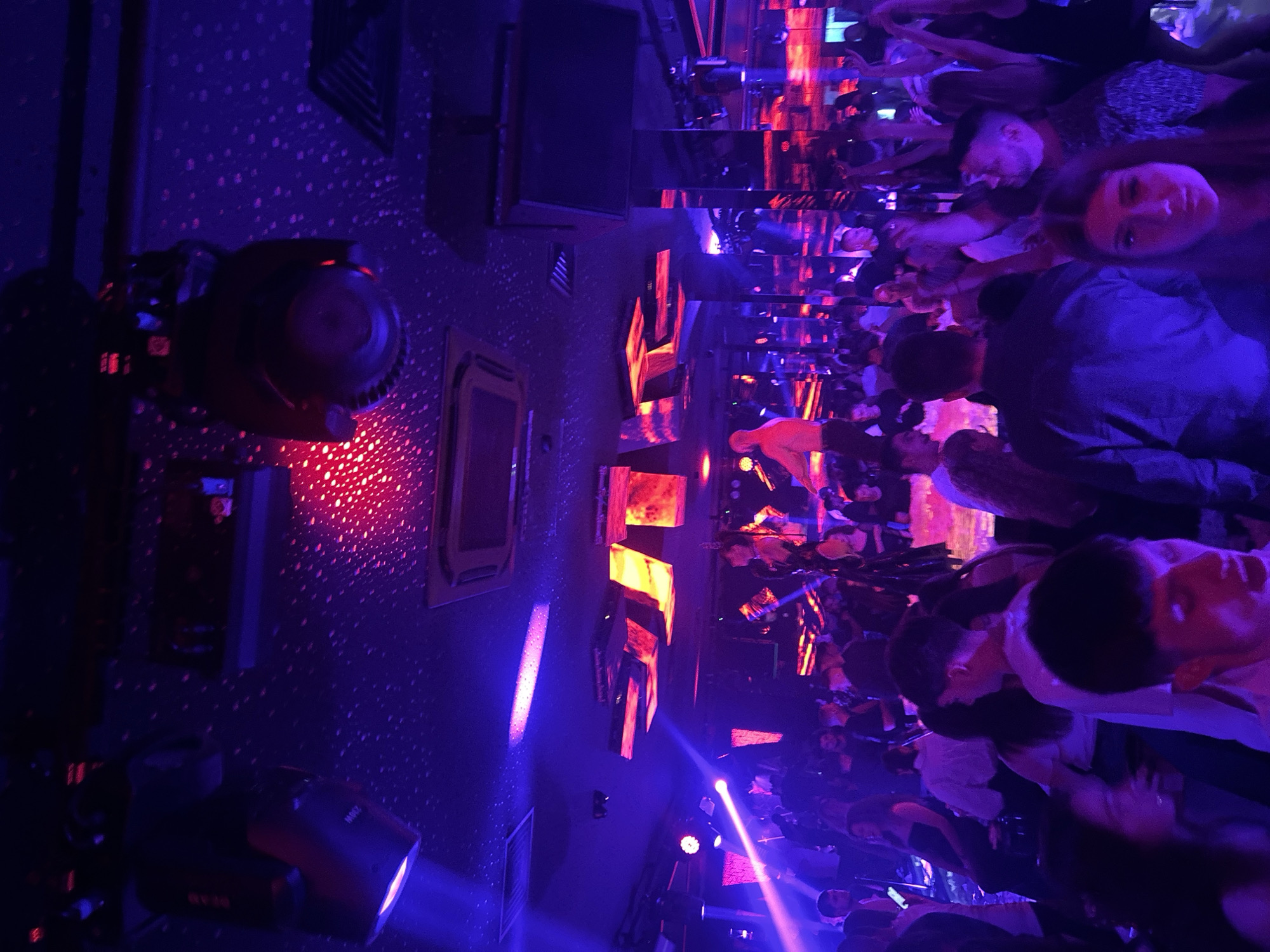 The image depicts a crowded nightclub scene, illuminated by blue and red lighting effects. People are standing close together, with some facing the camera. Mirrors line the walls, creating a fragmented reflection of the crowd and the lighting. In the foreground, there are stage lights mounted on the ceiling. The walls are decorated with rectangular frames. Based on context clues, it is likely that the picture was taken in a nightclub in the city of Sofia, Bulgaria.