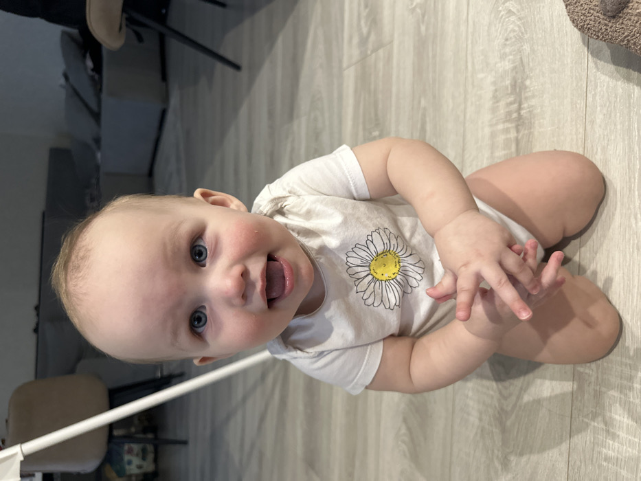 An infant with fair skin, light hair, and blue eyes is depicted kneeling on a light wood-patterned floor in an indoor residential setting in Chaikovsky, Russia. The infant wears a white short-sleeved onesie featuring a yellow-centered daisy graphic. The subject faces the camera directly, mouth slightly open, and hands clasped together at chest level. In the blurred background, a white vertical support structure is visible on the left, along with a light beige chair with dark legs, a grey modular sofa unit, and a dark-legged chair on the right. A textured, brown object is partially visible on the floor in the lower right corner.