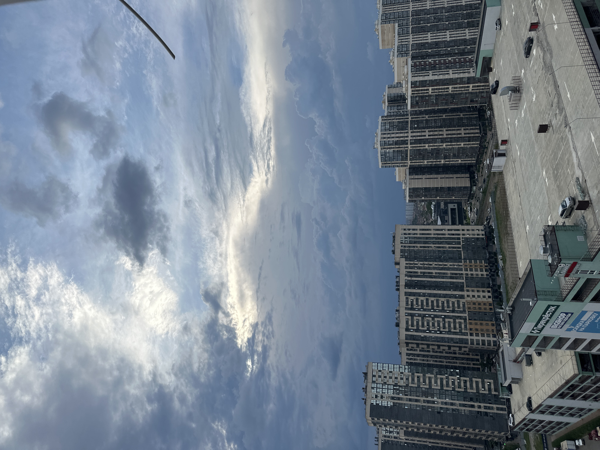 The image captures an aerial view of a residential complex in the city of Krasnodar, Russia, under a dramatic, overcast sky. The sky is filled with a variety of clouds, ranging from dark, heavy formations to lighter, wispy cirrus clouds, with a bright patch of sunlight breaking through in the upper left quadrant.
Dominating the foreground and midground are several tall, modern apartment buildings characterized by their uniform facade of windows and vertical architectural lines. These buildings appear to be relatively new. In front of the apartment buildings is a large, multi-level parking structure, likely part of the residential complex. The parking structure is mostly empty, with a few cars visible on its upper levels. One section of the parking structure has a green facade with Russian text that includes "Перекрёсток" (Perekrestok), a common supermarket chain in Russia.
The environment appears to be an urban setting, with other buildings and infrastructure visible in the distance, suggesting a densely populated area. The overall scene suggests a cloudy, possibly pre-storm or post-storm atmosphere. No people are clearly visible in the image.