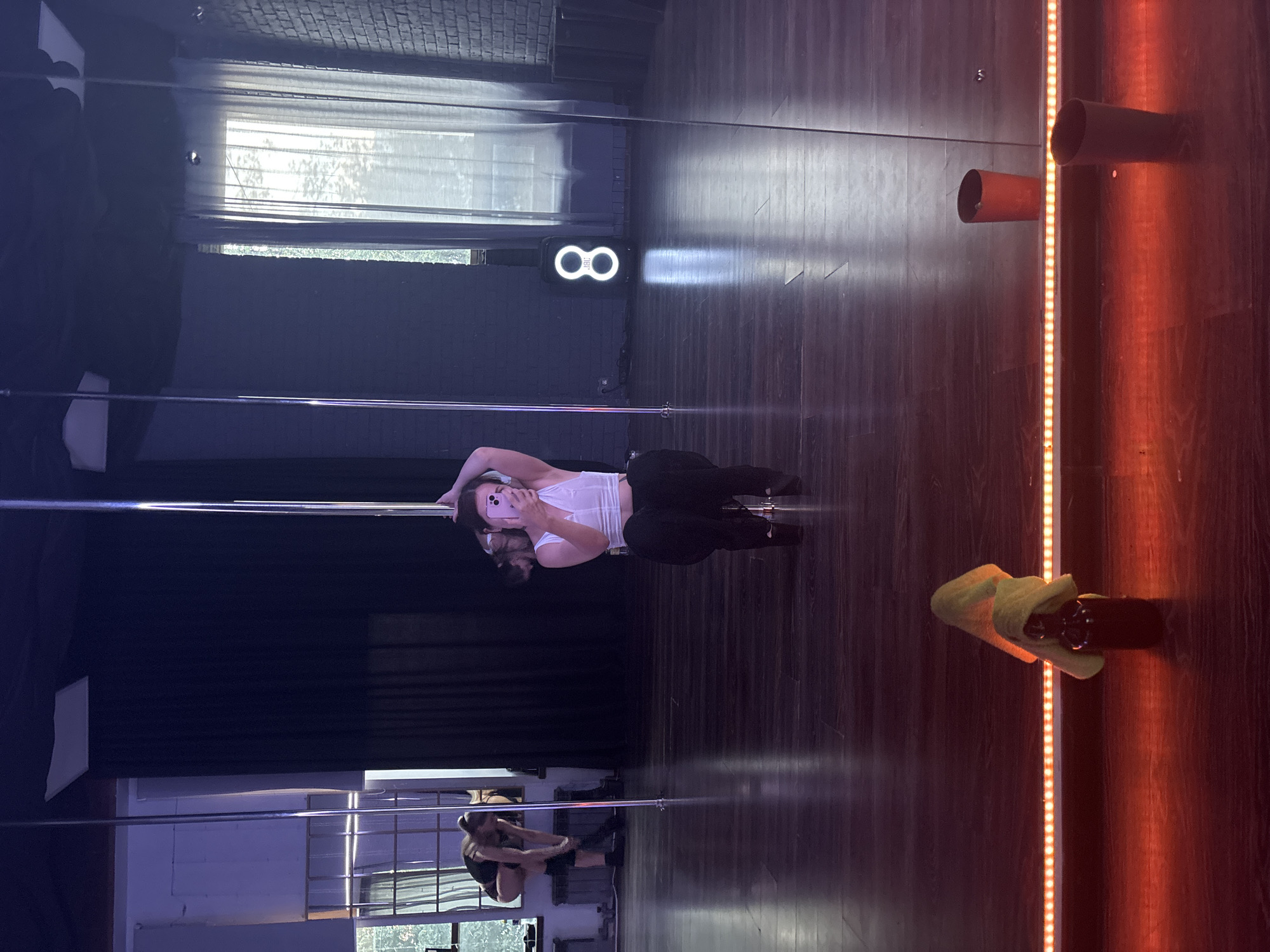 A young woman is taking a selfie in a dance studio. She is wearing a white crop top and black pants, and is holding a pink smartphone. The studio has polished wooden floors, mirrors, and a pole dancing apparatus. There are other people in the studio, including a woman tying her shoe in the background. The studio appears to be in the city of Naberezhnye Chelny, Russia.