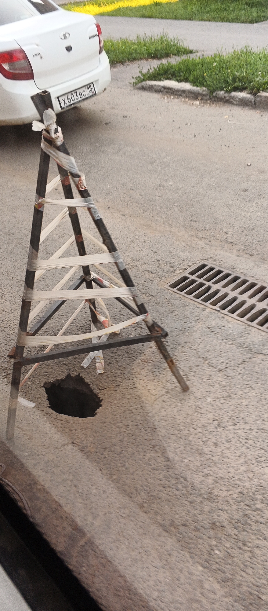A temporary barrier made of metal poles and reflective tape has been erected around a pothole on an asphalt road in Izhevsk, Russia. The barrier is triangular in shape, with the poles forming the legs of the triangle. The pothole is dark and appears to be deep. In the background, a white car with a license plate that reads "X603BC 18" is parked. A storm drain grate is visible to the right of the barrier. The ground is covered with asphalt and some patches of grass are growing along the side of the road. The scene appears to be a typical roadside hazard in a Russian city.