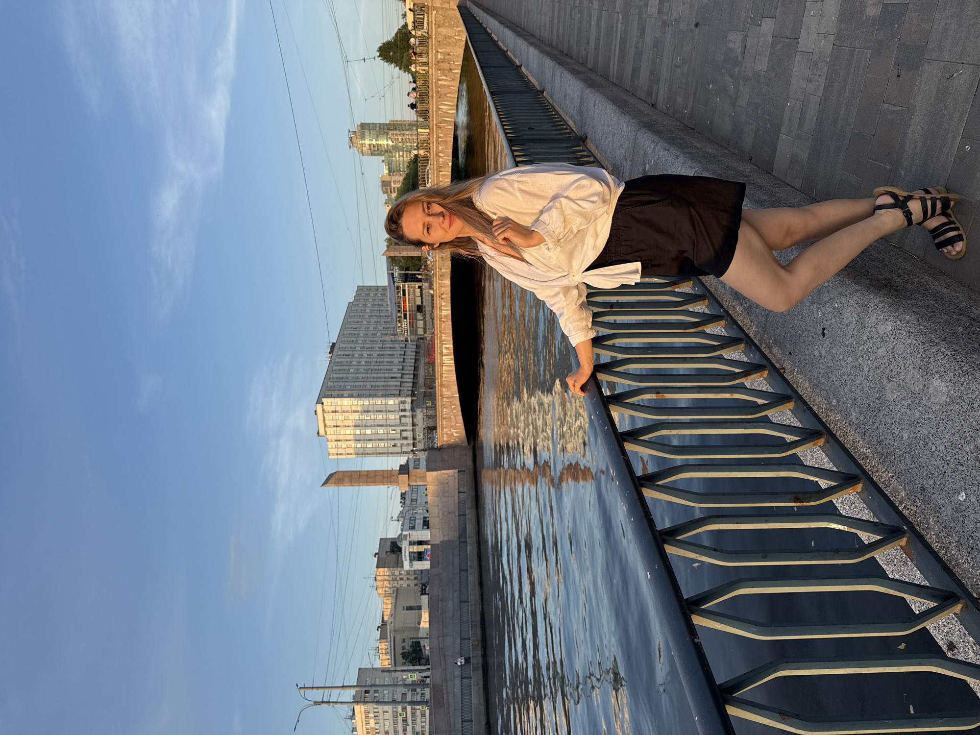 A woman is standing on a city embankment in Saint Petersburg, Russia, leaning against a black metal railing with a diamond pattern. She is wearing a white, loosely tied shirt and black shorts, with black sandals. Behind her, a wide river flows, reflecting the golden light of the setting sun. Across the river, there are buildings and a bridge. The sky is a clear blue with a few wispy clouds. The embankment is paved with light-colored stones.
