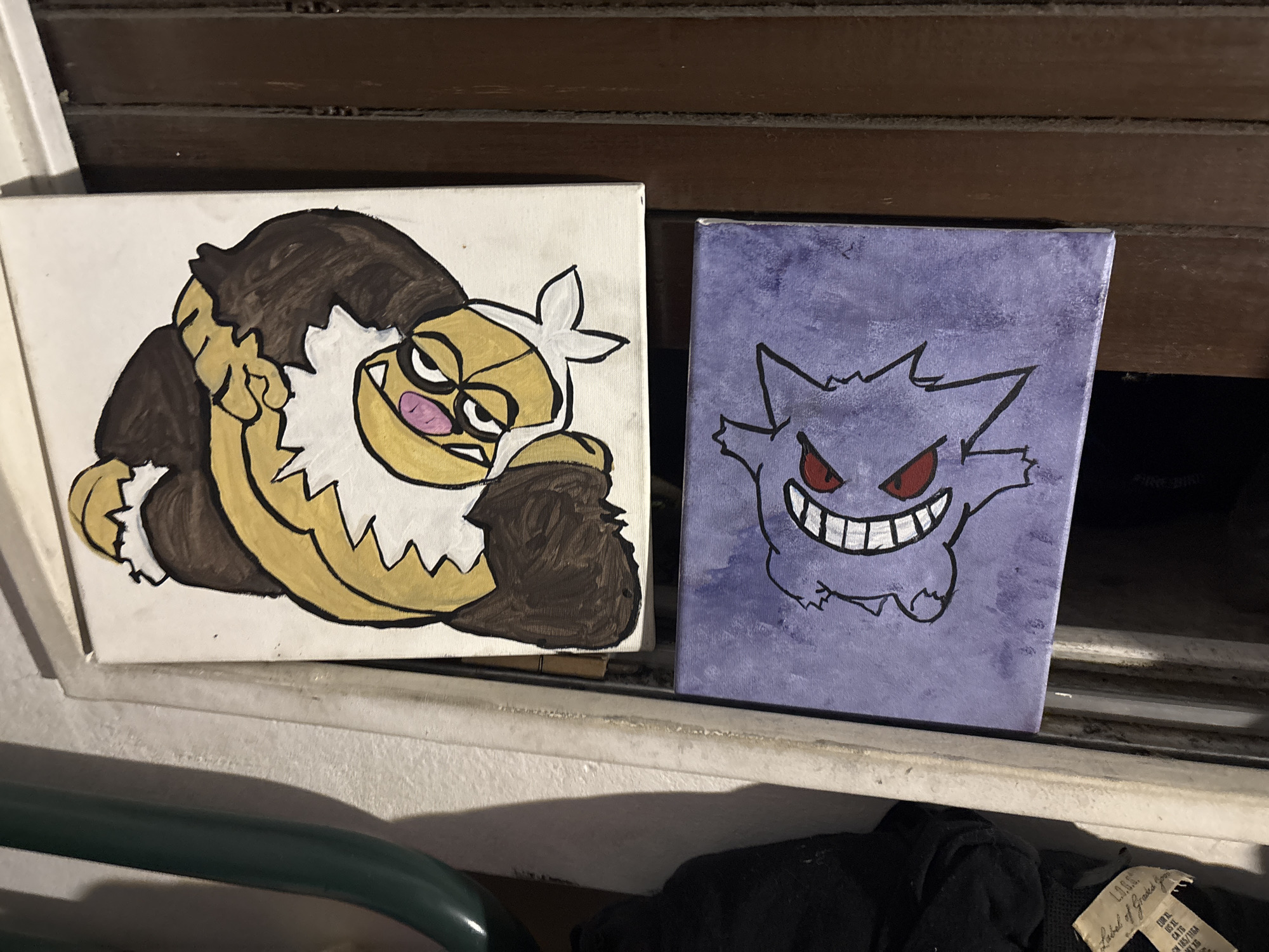 The image displays two small, vertically oriented painted canvases, positioned side-by-side on a light-colored ledge, likely a windowsill, within an indoor setting in Buenos Aires, Argentina.
The canvas on the left features a hand-painted character resembling a Pokémon, possibly Primeape or an evolution, set against a plain white background. This character has a bulky form with dark brown fur on its body and head, complemented by yellow skin on its face, chest, and limbs. It has white fluffy accents around its neck and wrists, and a white band on its head. The character's expression is angry or mischievous, with furrowed brows, yellow eyes, a pink nose, and a wide, toothy grin.
The canvas on the right depicts another Pokémon-like character, Gengar, against a textured purple background with visible brushstrokes. Gengar is presented as a shadowy, rounded figure with pointed ears and limbs, characterized by piercing red eyes and a wide, menacing smile revealing numerous white teeth.
In the background behind the canvases, a dark brown, horizontally slatted structure, possibly wooden blinds or shutters, is visible. The foreground includes a dark green metal railing and a piece of dark clothing with a light-colored tag. The tag on the clothing displays the text: "L.O.G.G.", "Label of Graded Goods", "EUR XL", "US XL", "CA TG", "MX TG", and "CN 185/110A". The lighting suggests an indoor environment. No specific human activity is shown.