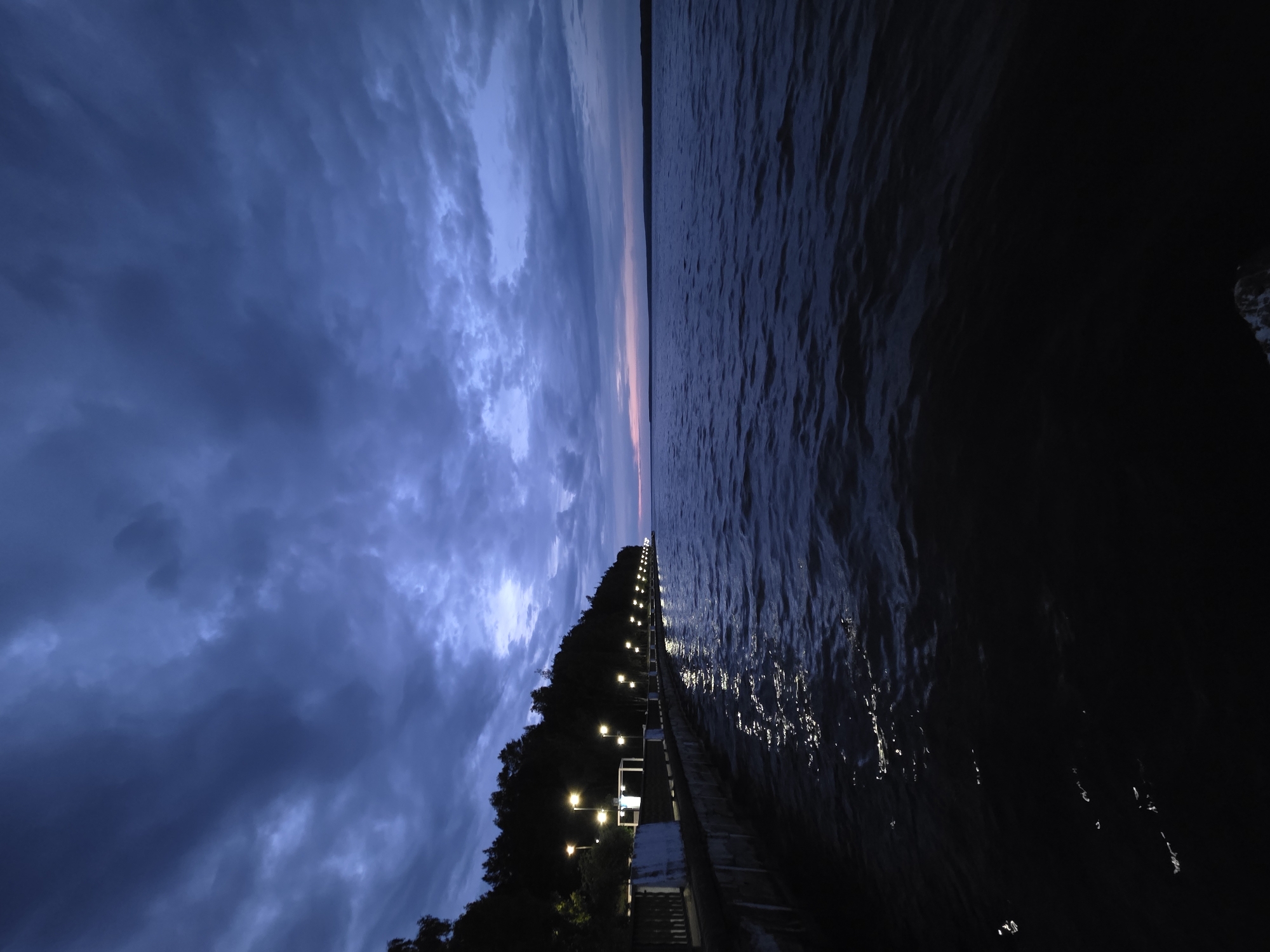 The image shows a long pier extending into a body of water, likely at dusk or twilight. The scene is located in Chaikovsky, Russia.
The pier is lined with evenly spaced lights, illuminating the walkway. To the left of the pier, trees are visible. On the right, the water reflects the light from the pier and the sky, creating a contrasting dark and reflective surface. The sky has a mix of dark, cloudy areas and a lighter band of color on the horizon, suggesting a sunset or sunrise.