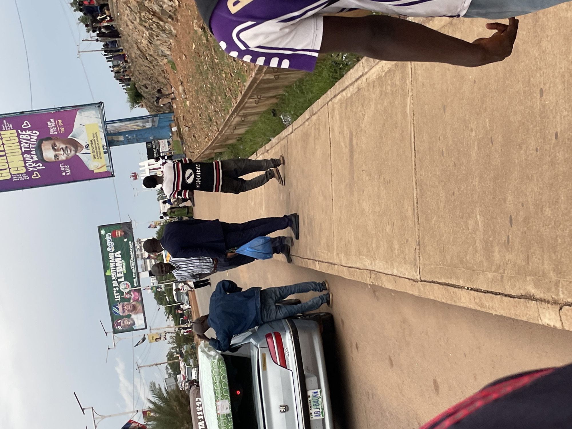 The image captures a street scene in Jos, Nigeria, with a prominent billboard in the background advertising "Gloryland Church." The billboard features a portrait of a young man and text that reads "YOUR TRYBE IS WAITING." To the left of this billboard, another large billboard displays multiple portraits, with text indicating "LET'S DO RUTTEWANG AGAIN" and "HON. EPHRAIM USMAN GAR."
In the foreground, several individuals are visible on a paved walkway. One man in a dark blue jacket and jeans is standing near the open trunk of a silver sedan, which has a mattress secured on top. He appears to be interacting with the car. Two other men are standing on the walkway, one in a striped shirt and the other in a dark suit. Further down the walkway, a person wearing a black and white striped long-sleeved shirt and dark pants walks away from the camera. On the right side of the image, the back of another person wearing a purple and white shirt is visible.
The environment is characterized by a slightly overcast sky and a dusty, reddish-brown ground typical of the region. Beyond the walkway, a grassy slope with some debris is visible. The overall impression is of a public area with a mix of urban advertising and everyday activities.