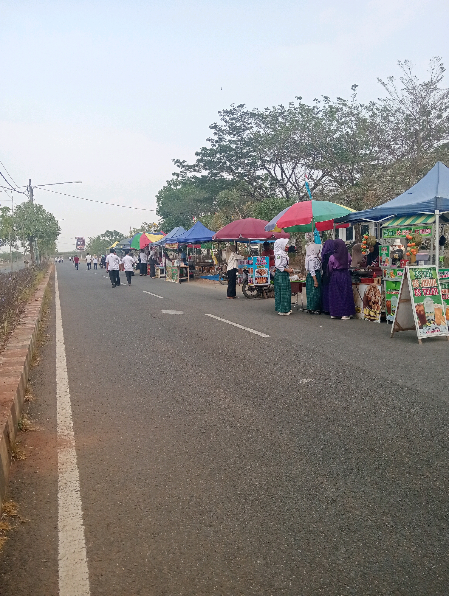 The image depicts a street scene in Sidoharjo, Indonesia, with a row of vendor stalls set up along the side of a paved road. Several people are visible, including vendors and customers. Some individuals appear to be students, wearing uniforms that include hijabs. The stalls are covered with colorful awnings and display various food and drinks, indicated by signs advertising items such as "Es Teh" (iced tea) and "Es Jeruk" (orange juice). In the background, trees and a utility pole are visible, suggesting an outdoor setting, possibly a market or a community event. The road has a white line marking the lane. The lighting suggests it is daytime, with a bright sky.