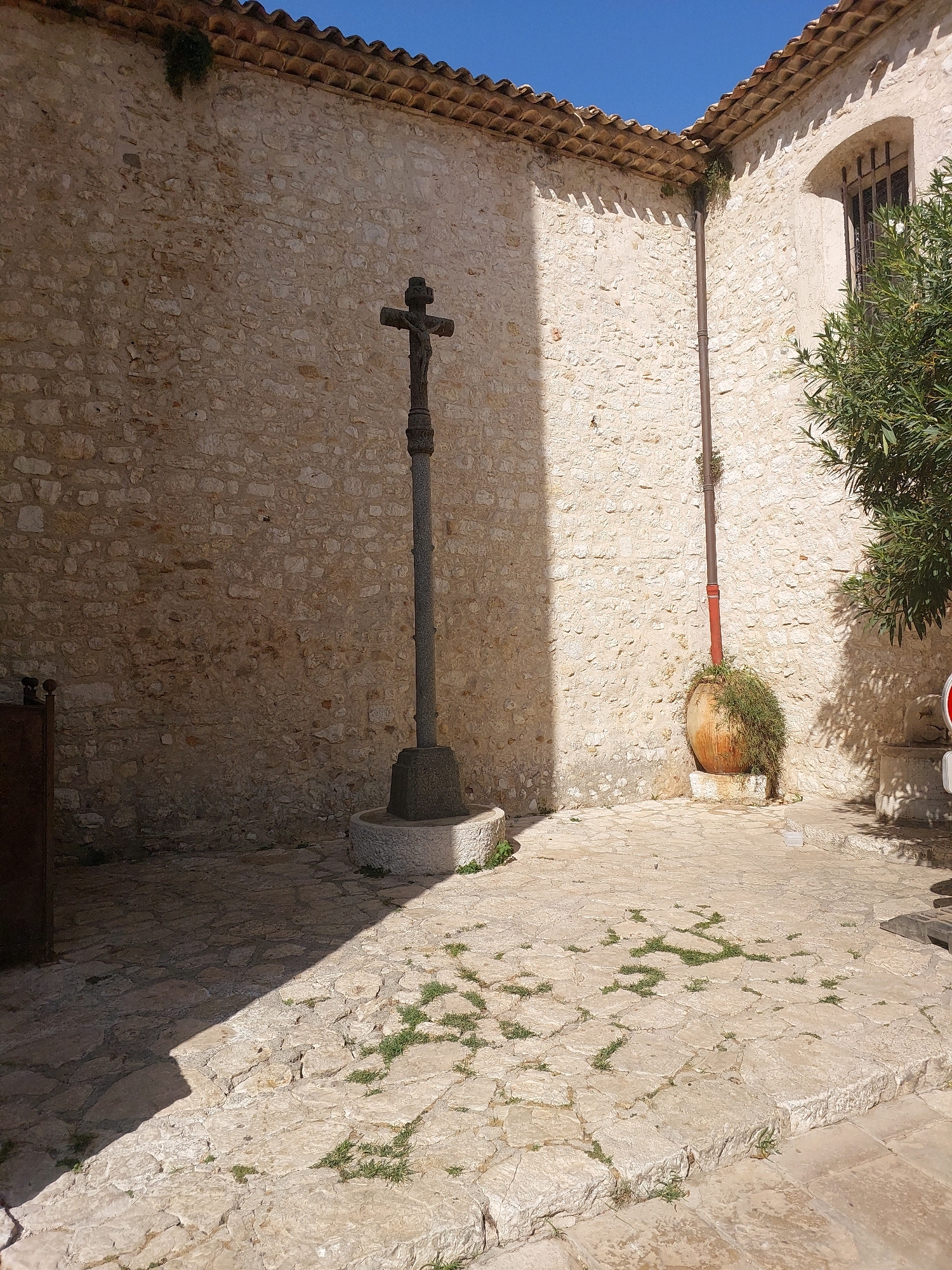 The image shows an outdoor stone courtyard in the village of Saint-Paul-de-Vence, France. The foreground features a stone path with scattered patches of grass growing between the paving stones. A large stone cross with a figure of Christ stands prominently in the middle of the courtyard, casting a shadow across the ground. Behind the cross, a textured stone wall forms the backdrop, with a terracotta tiled roofline visible at the top. To the right of the cross, a large, round terracotta pot overflows with greenery, and a downspout runs vertically along the wall. Further to the right, a window with barred panes is set into the stone wall, and a leafy green tree partially obscures the view. The scene is bathed in bright sunlight, creating strong contrasts between light and shadow.