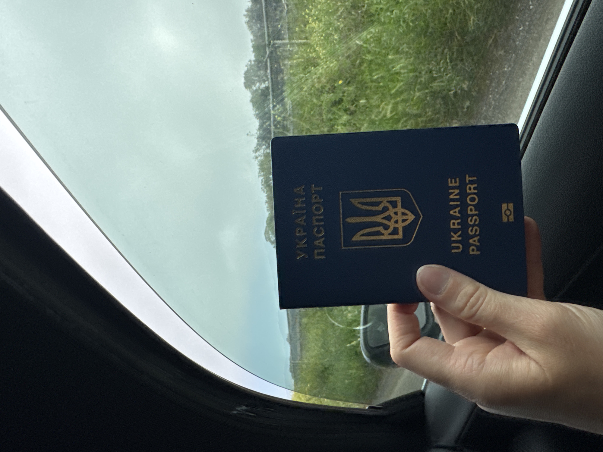 The image shows a hand holding a blue Ukrainian passport inside a car. The passport's cover displays the Ukrainian coat of arms and the words "УКРАЇНА ПАСПОРТ" and "UKRAINE PASSPORT" in gold lettering. The view through the car window reveals a blurred landscape with greenery and overcast sky. The photograph was possibly taken in the city of Cisterna di Latina, Italy.