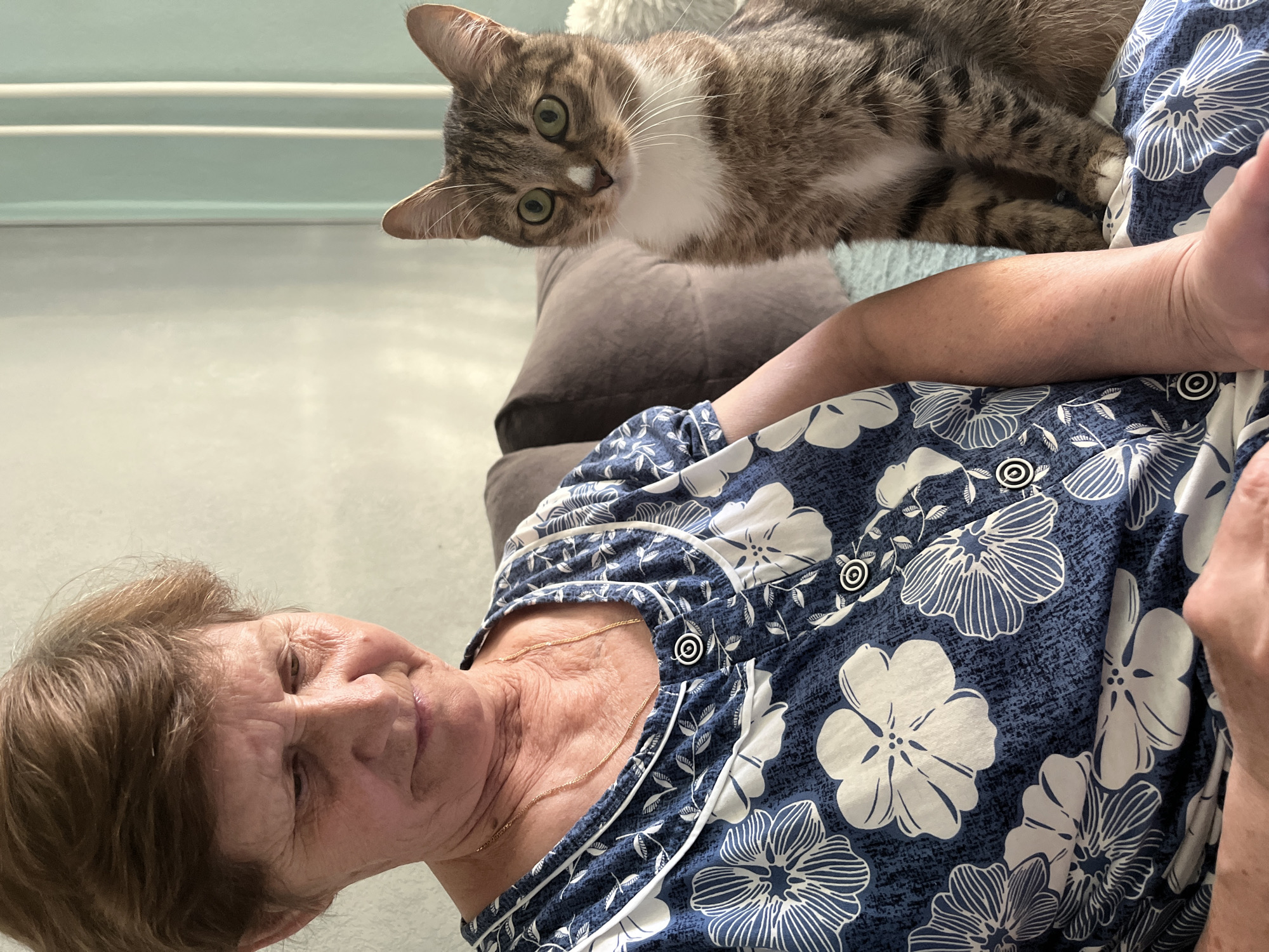 An elderly woman lies on a gray couch, wearing a blue and white floral-patterned garment with white-rimmed black buttons, a gold necklace resting at her neck. A brown tabby cat with white markings on its chest and face lies on top of the couch, looking downward at the woman. The background is a neutral gray wall. There is no evidence to identify this location as Izhevsk, Russia.