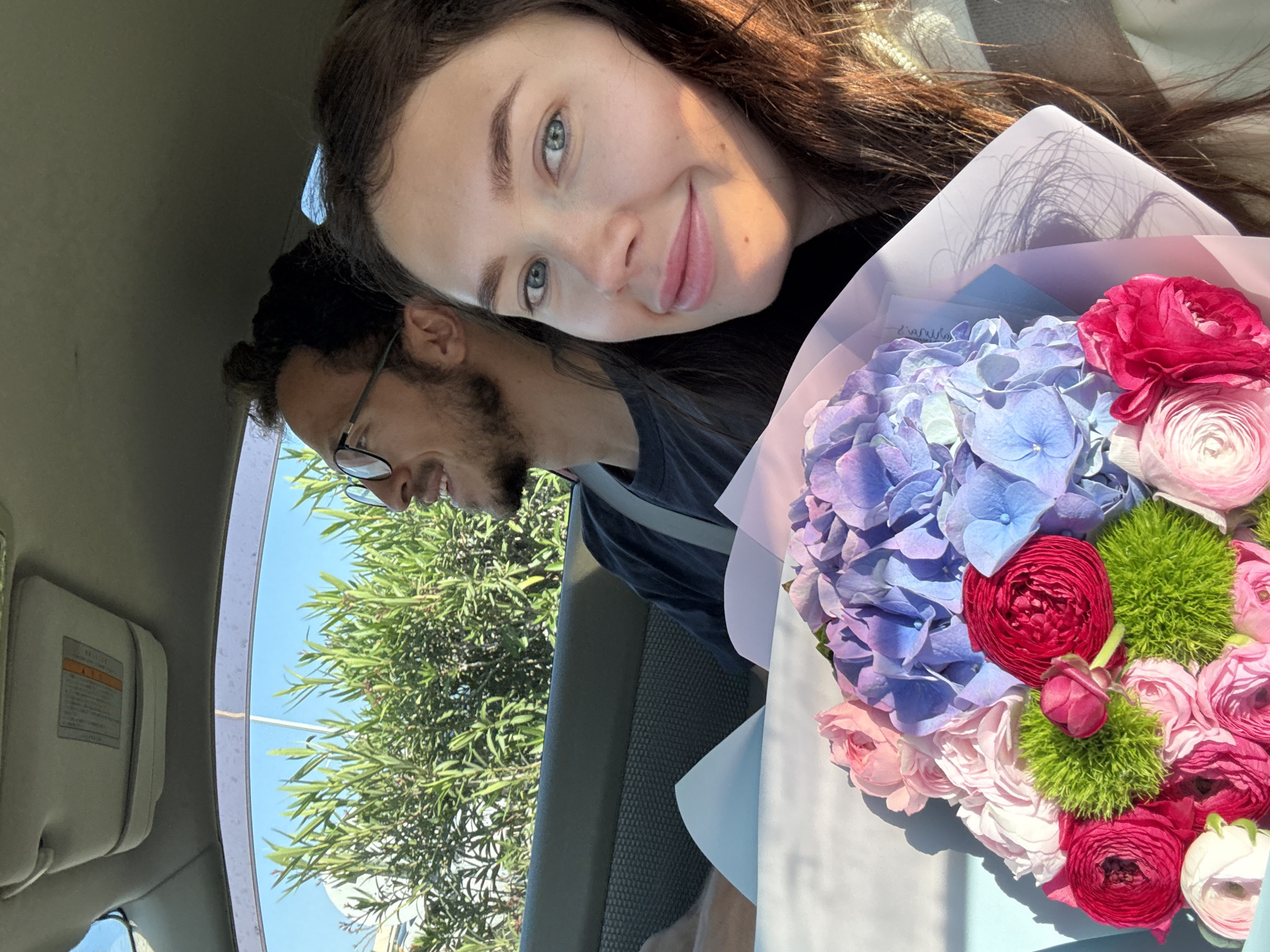 The image, rotated 90 degrees clockwise, captures a smiling couple inside a car in Dromolaxia - Meneou Municipality, Cyprus, on a bright, sunny day. A woman with long dark hair and blue eyes, illuminated by sunlight, smiles directly at the camera. She holds a large, colorful bouquet of flowers. Behind her and slightly to her side, a man with a beard and glasses is visible in profile, also smiling.
The vibrant bouquet features a mix of blue and purple hydrangeas, deep pink and red ranunculus, soft pink roses, and distinctive green spherical flowers, all wrapped in layers of light pink and light blue paper. A small, light-colored card with faint, partially illegible handwriting is tucked into the bouquet.
The setting is a car interior, with a beige sun visor and headliner visible on the left side of the frame (when viewed upright). Through the car windows, a clear blue sky and lush green foliage of trees are visible, indicating pleasant outdoor weather. The overall mood is happy and joyful. On the car's sun visor, there is a label with Japanese text.