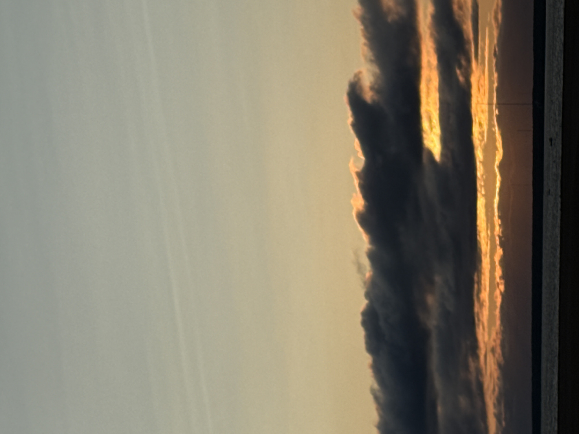 The image depicts a dusky sky over a body of water, likely a bay or river, in Saint Petersburg, Russia. The sky is a gradient of soft blues and yellows, with wispy clouds tinged with orange from the setting sun. A distinct band of darker clouds, heavily shadowed, stretches across the middle of the frame, with a sliver of bright orange sunlight breaking through it. Below the clouds, a dark horizontal line represents the distant shoreline, and the water is a deep grey. The overall impression is of a serene, perhaps cool, evening by the water. There are no people or specific landmarks visible to pinpoint a precise location within Saint Petersburg.