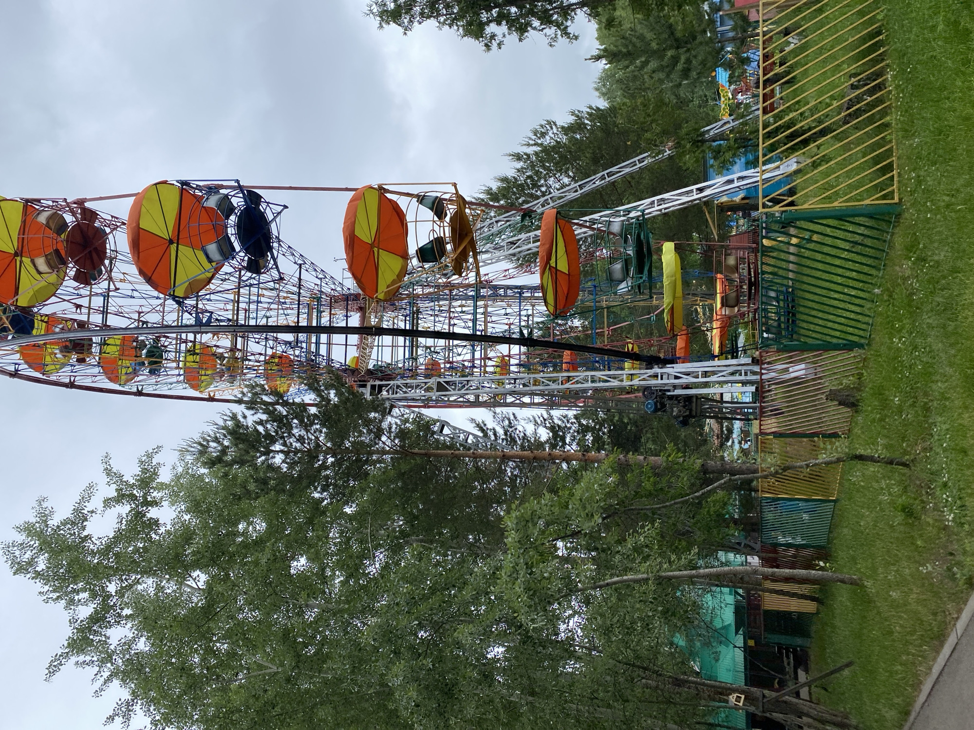 This image presents a Ferris wheel within a park setting in Chaikovsky, Russia, under an overcast sky. The Ferris wheel, occupying the right side of the frame, features a robust metal structure of interconnected beams painted in multiple colors, including red, blue, and yellow. It is equipped with numerous gondolas, each topped with a two-toned umbrella-like canopy, predominantly orange and yellow, with some appearing solid green, yellow, or orange. Exposed seats are visible within some of the gondolas. A white, ladder-like support structure is visible running diagonally up the main frame on the right.
The left and central foreground are densely covered with green foliage, including deciduous trees with light-colored bark and several coniferous trees. A narrow asphalt or concrete pathway is visible along the bottom-left edge. A multi-colored picket fence, composed of sections painted dark green, yellow, maroon, light blue, and lime green, with diagonally oriented pickets, extends across the middle ground. Behind this fence, a building with a green corrugated roof is partially visible on the far left, alongside other obscured structures and additional trees. The sky is uniformly gray and cloudy. No individuals or animals are discernible within the scene.