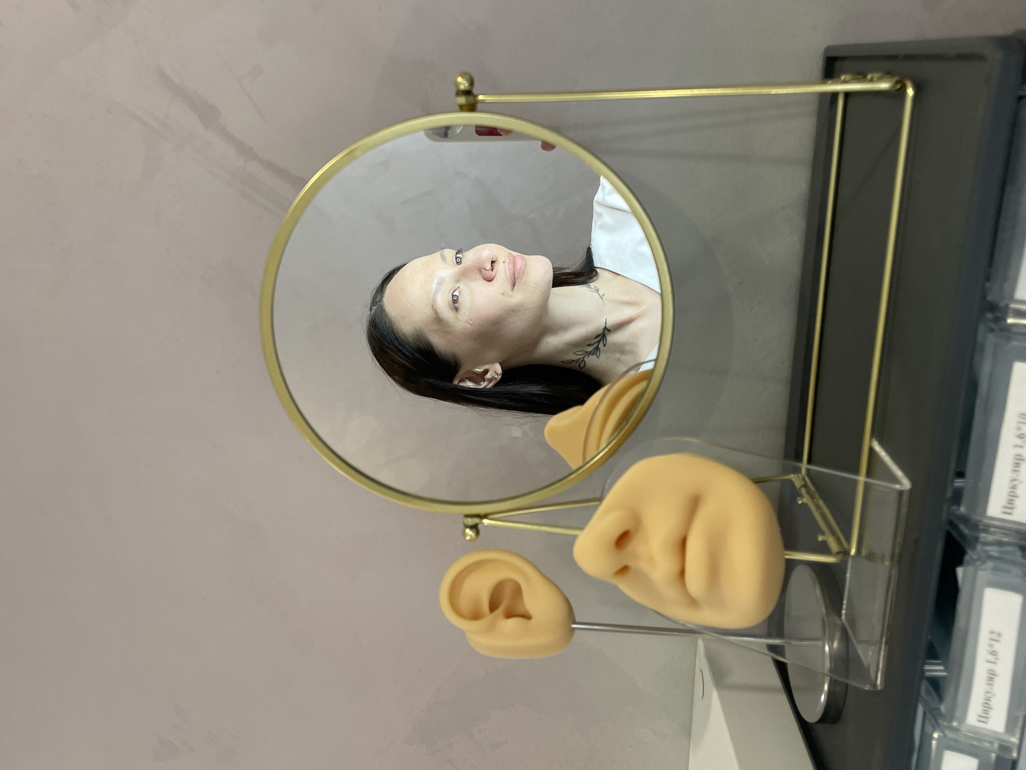 The image shows a person's reflection in a round, gold-framed mirror. The mirror is standing on a surface. In the foreground, there is a prosthetic nose and ear. The mirror is set up on what appears to be a display stand next to what looks like boxes with labels in Russian, including "Циркуляр 1,6*12". The setting seems to be an indoor location, potentially a store or studio in Izhevsk, Russia, given the text in Russian. The individual reflected in the mirror has dark hair and a visible tattoo on the neck.