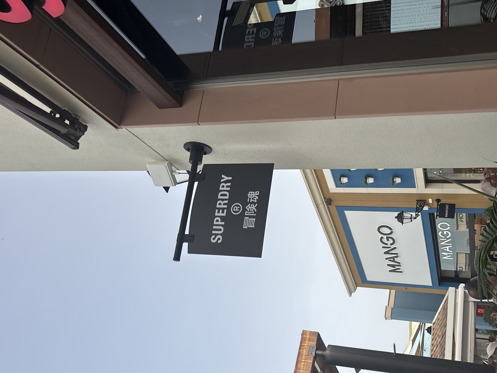 The image shows an outdoor scene with a clear sky. A black sign with white lettering hangs from a pole, displaying "SUPERDRY" and Chinese characters. To the right, a building has a pink facade and a large window reflecting the sky and a sign that reads "SUPERDRY" in reverse. Further to the left, another building features a white sign with "MANGO" in black letters, and a smaller "MANGO" sign is visible to its right. There is a black lamppost to the right of the "MANGO" sign. The overall setting appears to be a commercial area, possibly a shopping outlet, in Orihuela, Spain, given the presence of recognizable retail brands. The image is taken from a slightly upward angle.