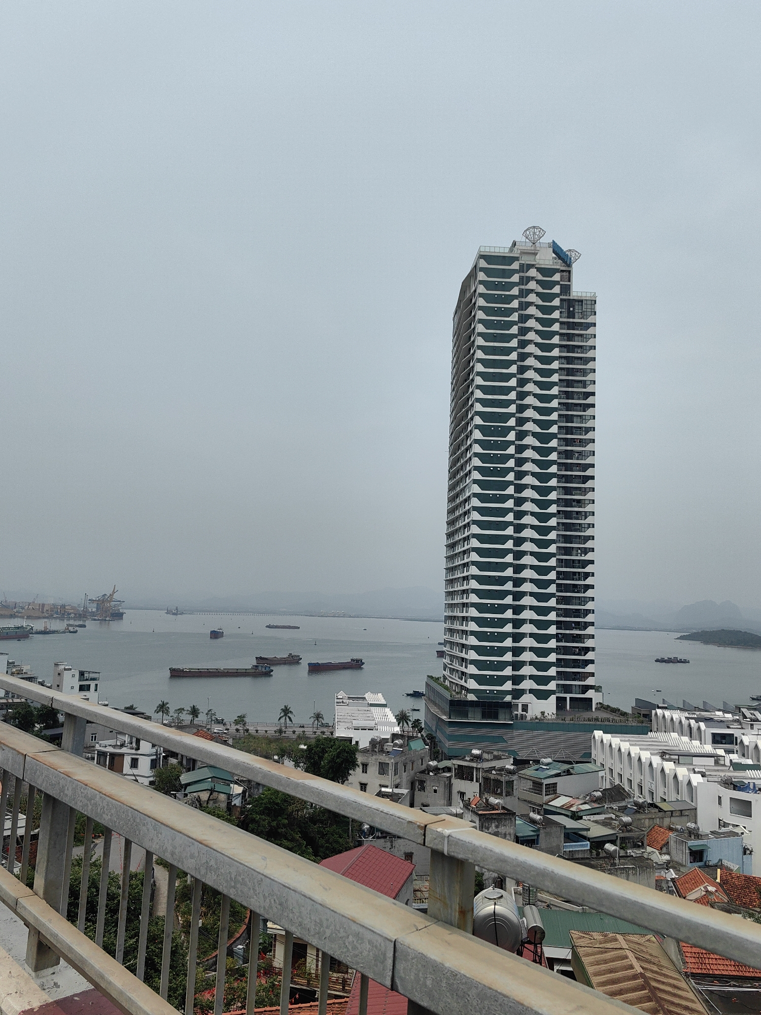 The image depicts a cityscape in Hong Gai Ward, Vietnam, featuring a prominent modern high-rise building with a distinct white and green facade. The building is located near the waterfront. In the background, a body of water is visible with several ships, and a distant shoreline. The foreground includes a metallic railing, partially obscuring the view of rooftops of smaller buildings and houses. The sky is overcast, giving the scene a muted color palette. The environment appears urban, with a mix of residential and possibly commercial structures. No people are clearly visible in the image.