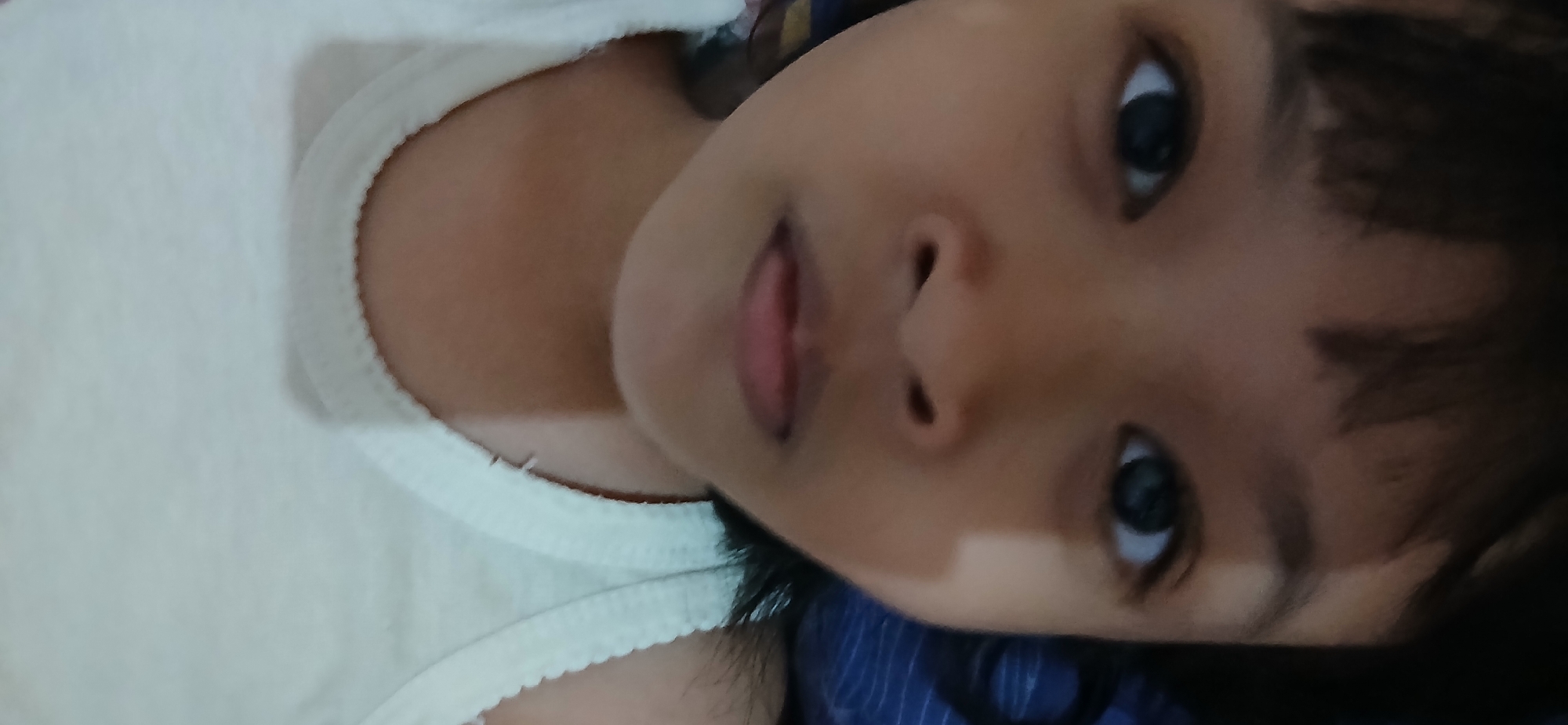 The image is an upside-down, close-up selfie of a young child's face. The child is lying down and looking at the camera. The child's head is resting on a blue and white striped surface, possibly a pillow or blanket. The child is wearing a white tank top with a ribbed neckline. The background is blurred, but appears to be indoors. The image was taken at the child's home in Samarinda, Indonesia.