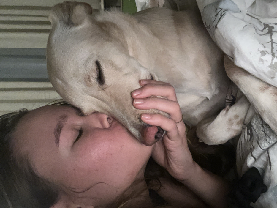 A woman with closed eyes embraces a light-colored Labrador Retriever in bed. Her cheek rests against the dog's head, and her hand gently cradles its face near the nose. The dog also has closed eyes. They seem comfortable and relaxed. The bedding around them is light-colored with a patterned design.