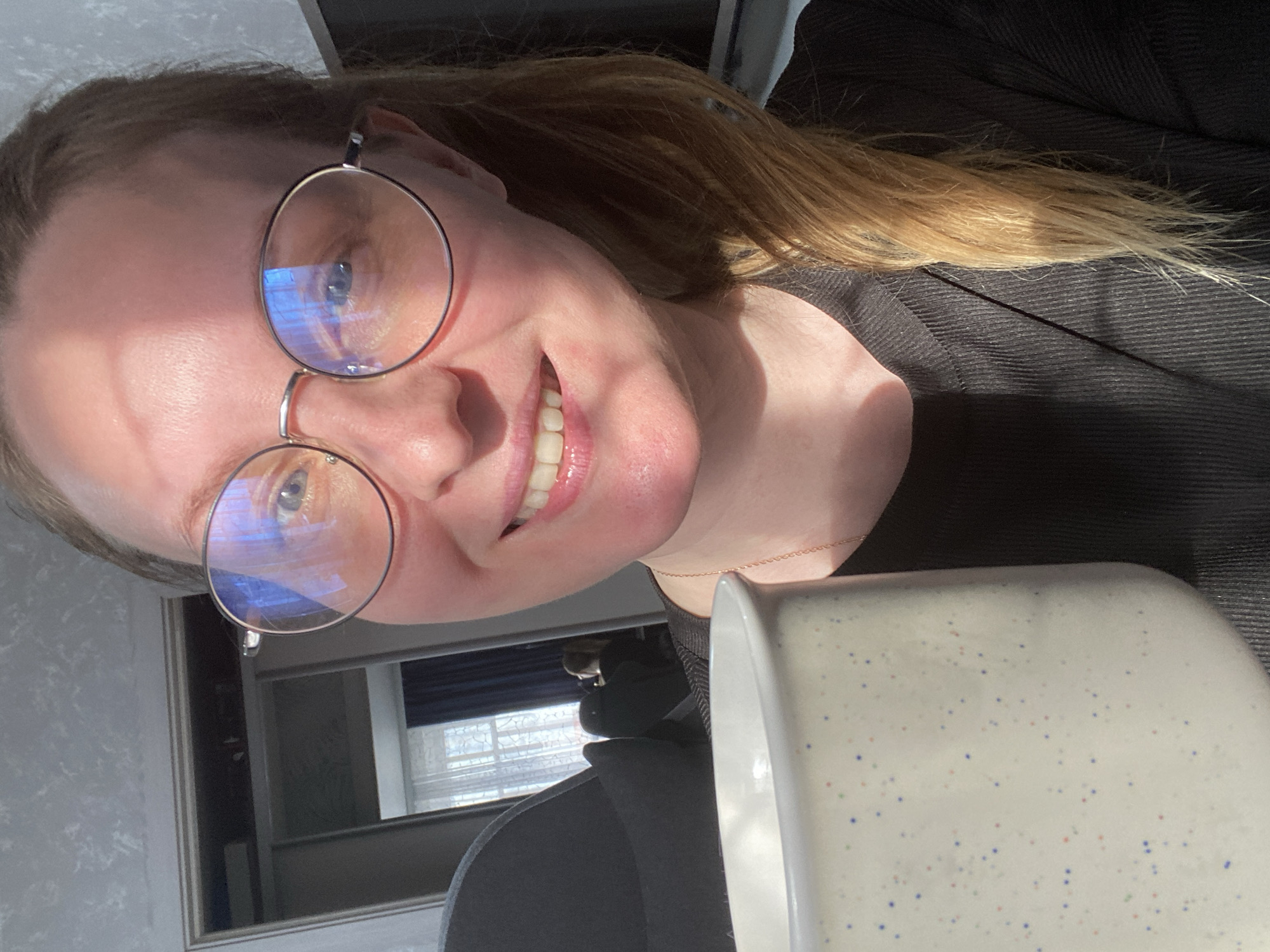 The image shows a woman with blonde hair pulled back, wearing round glasses and a black shirt, smiling at the camera. She is holding a white mug with blue and red speckles. Behind her is a window with blue curtains. The location of the photo is difficult to ascertain without further information, but there's no definitive evidence in the image to confirm it's taken in Noviy, Russia.