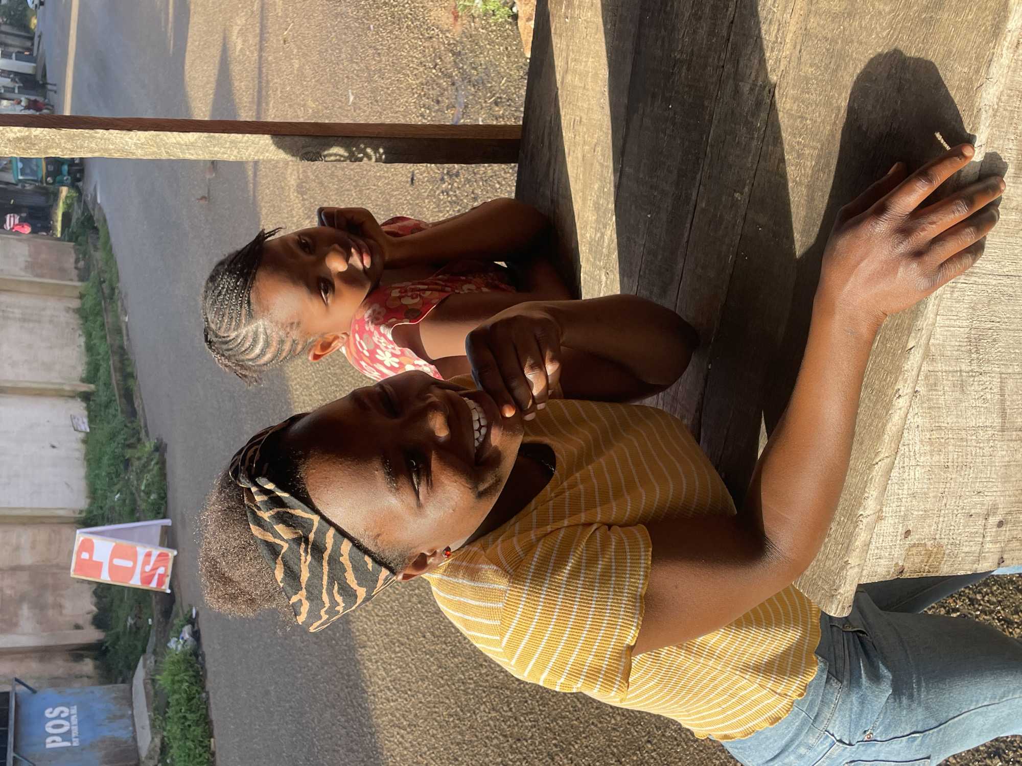 A medium shot shows two young Black females in the foreground, with a street scene and building in the background. The female on the left, closer to the camera, is leaning on a wooden surface with her left arm. She is wearing a yellow and white striped t-shirt and blue jeans, with a black and tan patterned headscarf tied around her hair. She is smiling and looking to her right. The second female is positioned behind her, to the right. She is wearing a pink floral-patterned dress and is resting her chin on her hands, looking towards the right edge of the frame. The wooden surface appears to be part of a table or counter. In the background, there is a street with a few people and a green tricycle. A white A-frame sign with red lettering that reads "POS" is visible to the left. A blue utility box with "POS" written on it is also present. The lighting suggests it is daytime, with shadows cast across the scene. The overall environment appears to be an outdoor, possibly commercial, area in Jos, Nigeria.