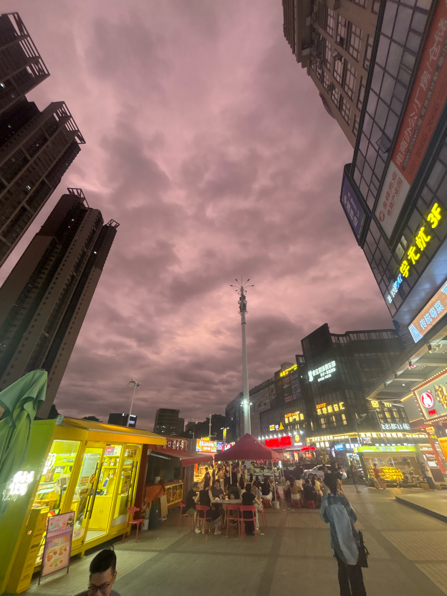This is a low-angle, wide shot of a city street in Nanhai District, China, at dusk. The sky is filled with a dusky pink and purple cloudy atmosphere. Towering residential and commercial buildings dominate the background, their windows reflecting the fading light. In the foreground, there is a bustling street scene with food stalls and outdoor seating. A bright yellow food stall is prominently featured on the left, illuminated from within, displaying various food items. Several people are seated at tables outside, some eating and others conversing. A person in the foreground is walking away from the camera, their back to the viewer. Neon signs illuminate some of the storefronts, creating a vibrant and active nighttime atmosphere.