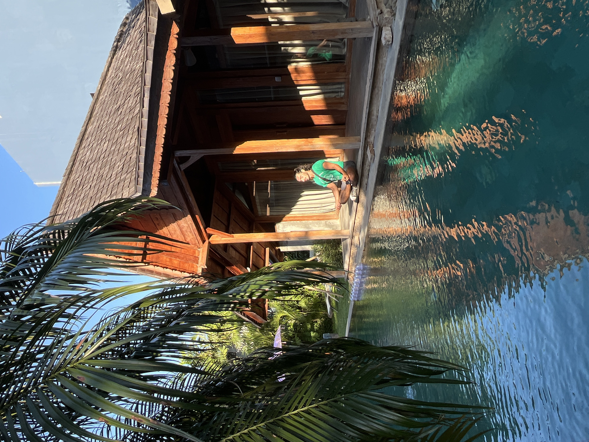 In Kuta, Indonesia, a man is meditating in a lotus position on the edge of a swimming pool, situated in front of a wooden building with a thatched roof. The sun casts long shadows across the scene, highlighting the tropical foliage and the rippling water of the pool. The man, wearing a green t-shirt and dark pants, has his eyes closed, seemingly engrossed in his practice. The surrounding environment suggests a tranquil and serene atmosphere, typical of a resort or private residence in Bali. The wooden structure, possibly a villa or bungalow, features open-air verandas and large windows, reflecting the vibrant greenery and the bright Indonesian sky. The pool's surface is a mosaic of light and shadow, reflecting the architectural details and the lush vegetation.