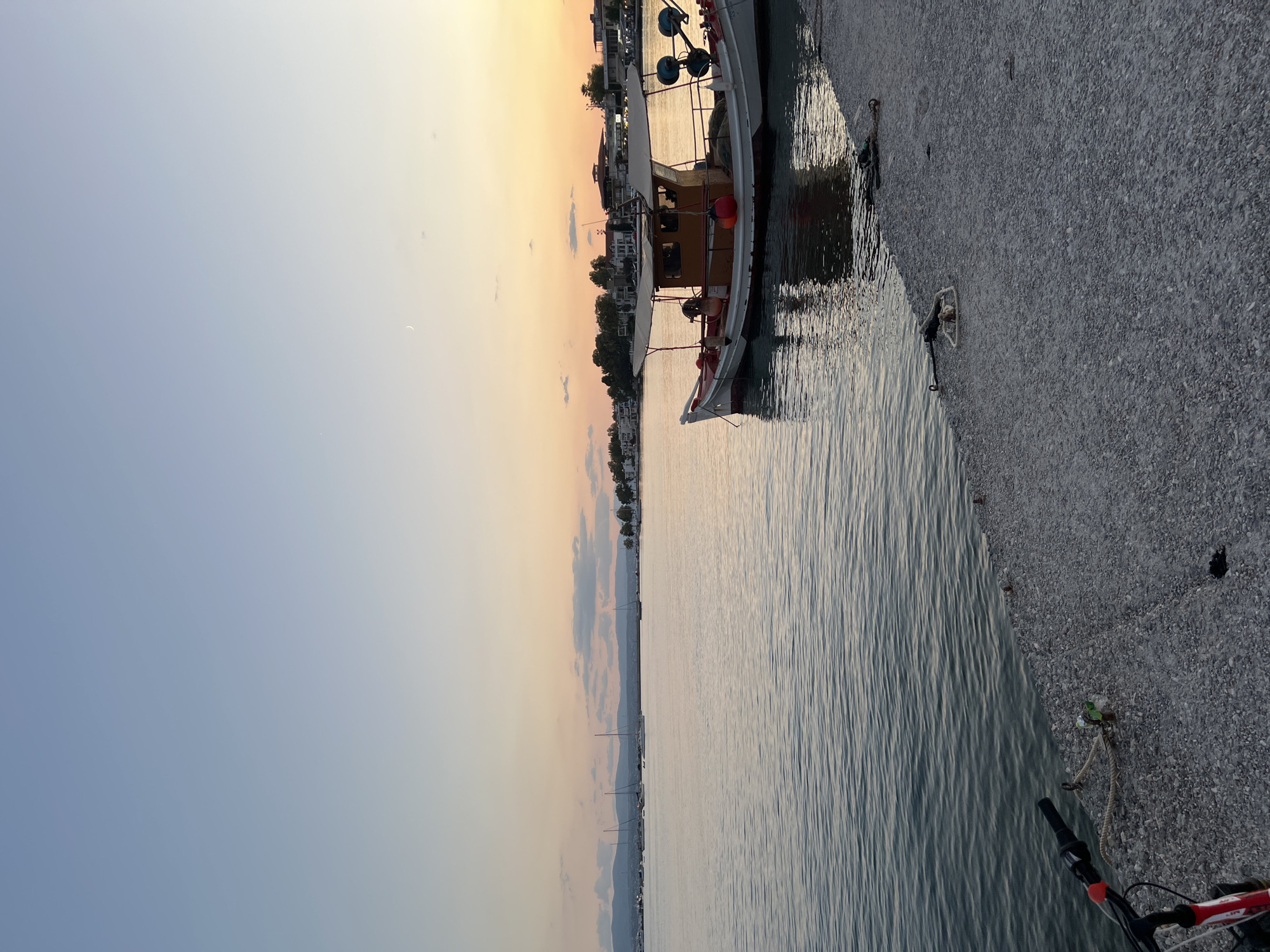 In the city of Eretria Municipal Unit, Greece, a fishing boat is moored at a harbor during sunset. The sky displays a gradient of colors from light blue to orange, with a sliver of a crescent moon visible. The water is calm, reflecting the sky and the boat. The boat is white with red trim, featuring a wooden cabin and a canvas covering. Several red buoys are attached to the boat. In the background, a town with buildings and trees lines the coast, with mountains in the distance. The foreground shows a concrete pier with a bicycle parked on the left side. Ropes and mooring cleats are visible on the pier. The scene is serene and tranquil.