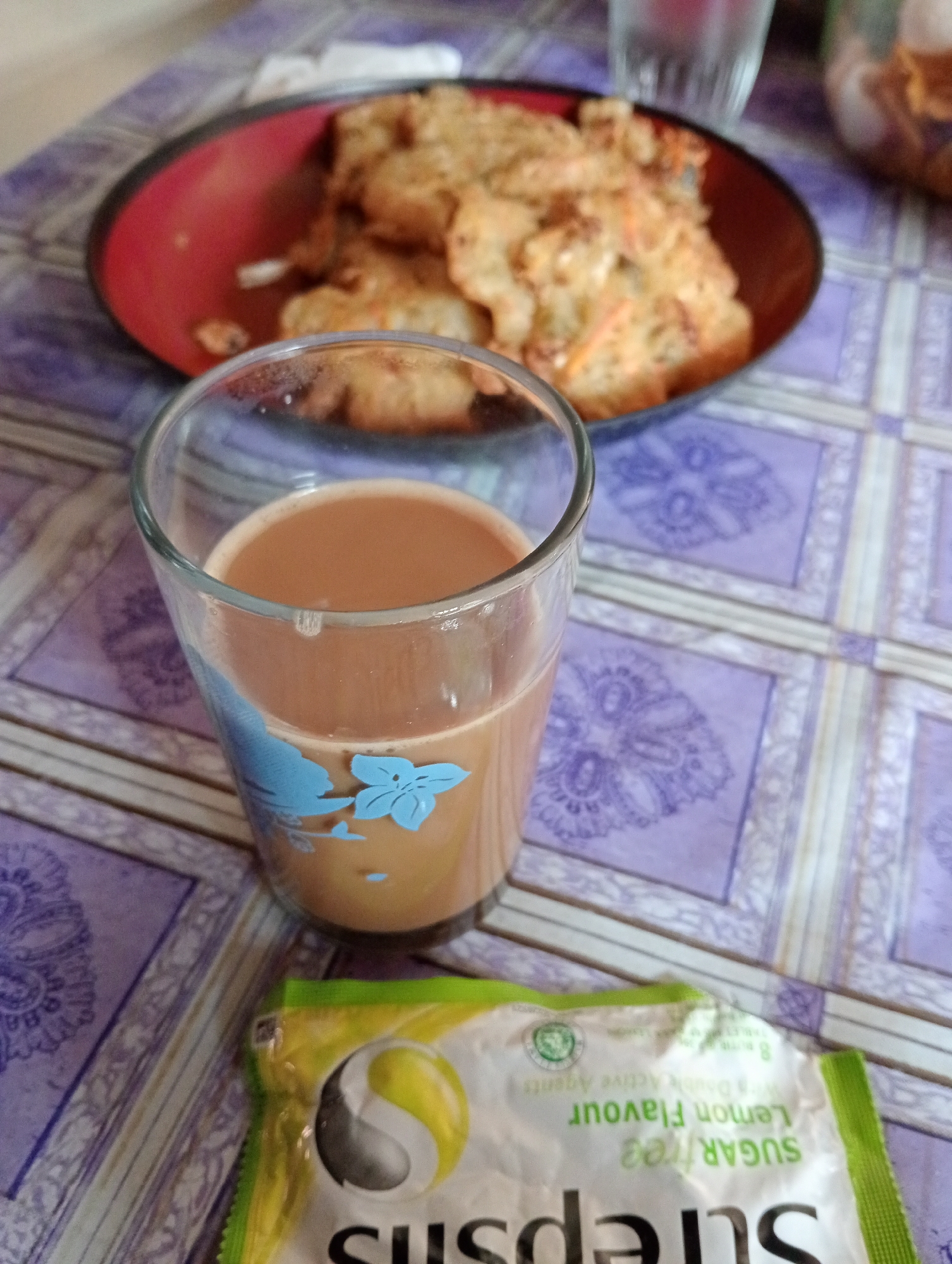 In Krajan Kidul, Indonesia, an overhead shot captures a breakfast scene on a purple and white checkered tablecloth. A red-rimmed plate holds a stack of golden-brown, fried food, possibly fritters or pancakes, with visible flecks of orange, likely carrots. In front of the plate, a clear glass contains a light brown beverage, likely tea or coffee, with a faint blue floral design on the exterior. To the right of the glass, a portion of a white and green foil packet is visible, displaying text that reads "SUGARS" and "Lemon Flavour". The setting appears to be an indoor dining area, with no people or animals visible.