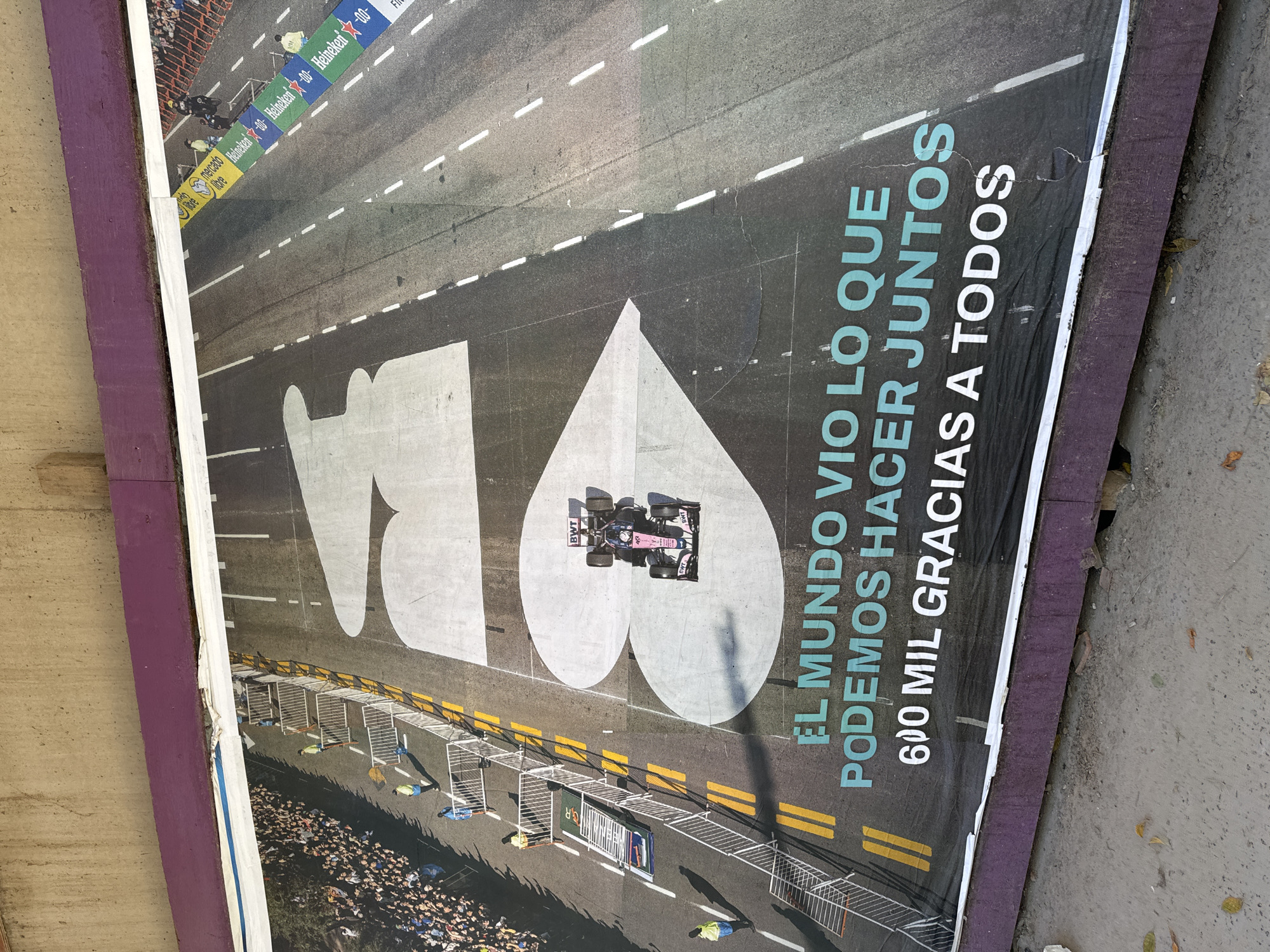 The image shows a poster related to Formula 1 racing in Buenos Aires, Argentina. The poster, likely advertising the event, depicts a Formula 1 race car driving over a racing circuit. The track has a large "AR" painted on it, presumably standing for Argentina. There are crowds lining the track behind safety barriers. The poster has text in Spanish: "EL MUNDO VIO LO QUE PODEMOS HACER JUNTOS 600 MIL GRACIAS A TODOS." Advertising from Heineken is visible at the top of the poster, and BWT is printed on the race car, likely as part of the livery. The poster is torn and damaged. It leans against a wall on a sidewalk.