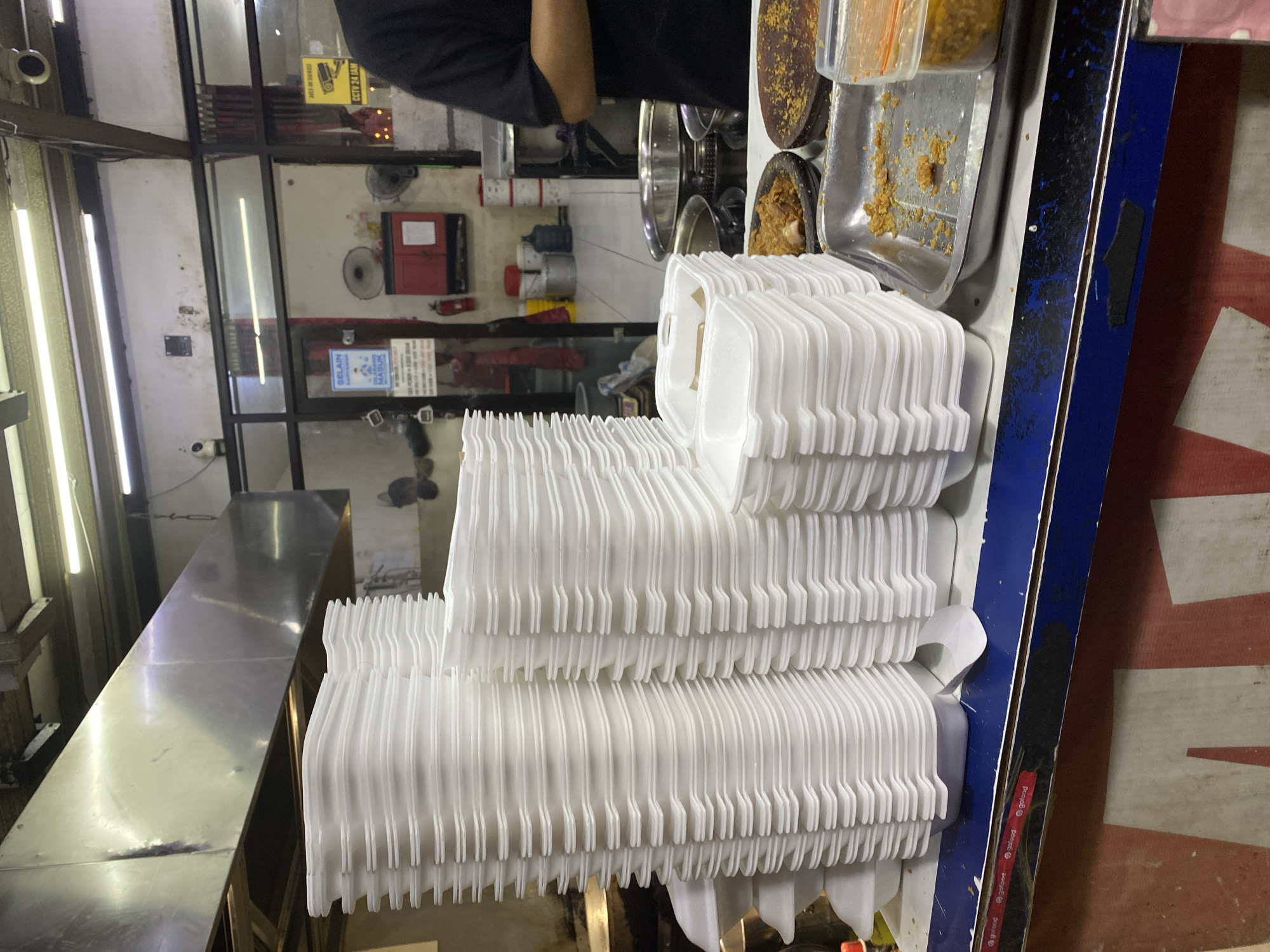 The image depicts a food stall in East Jakarta, Indonesia, with a focus on stacked white styrofoam food containers. These containers are piled high on a blue counter. To the right of the containers, there is a metal tray holding what appears to be fried food, and a clear plastic container with a yellow, granular substance. In the background, a person in a black t-shirt is partially visible, standing behind the counter. The stall appears to be indoors, with industrial-style lighting and ventilation hoods visible overhead. A sign with "CCTV 24 JAM" (CCTV 24 Hours) is mounted on the wall, indicating a security measure. Various other items, including a fire extinguisher and a red cabinet, are also present in the background, contributing to the utilitarian atmosphere of a busy food establishment.