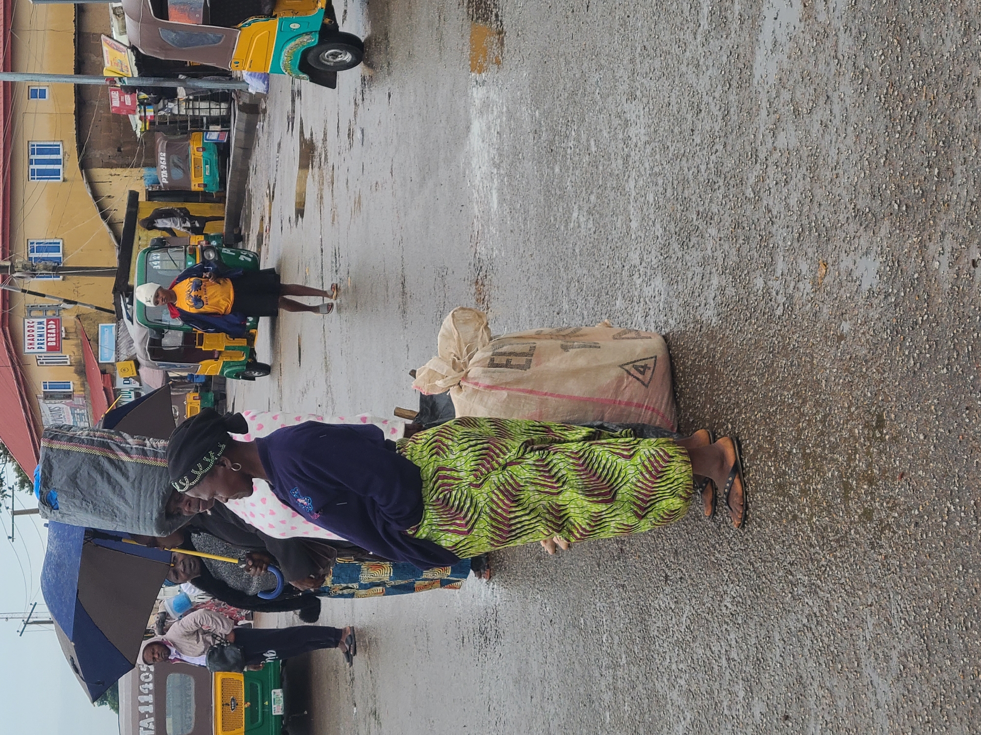 In Gura Top, Nigeria, a woman in a purple long-sleeved shirt and a patterned green and brown skirt stands on a wet, gray road next to a large beige sack. She wears black sandals. Another woman carrying a gray bag on her head stands under an umbrella beside her. Several auto rickshaws are parked or moving in the background, and other people are visible. Buildings, some painted yellow and red, line the street. The sky is overcast, suggesting it has recently rained.