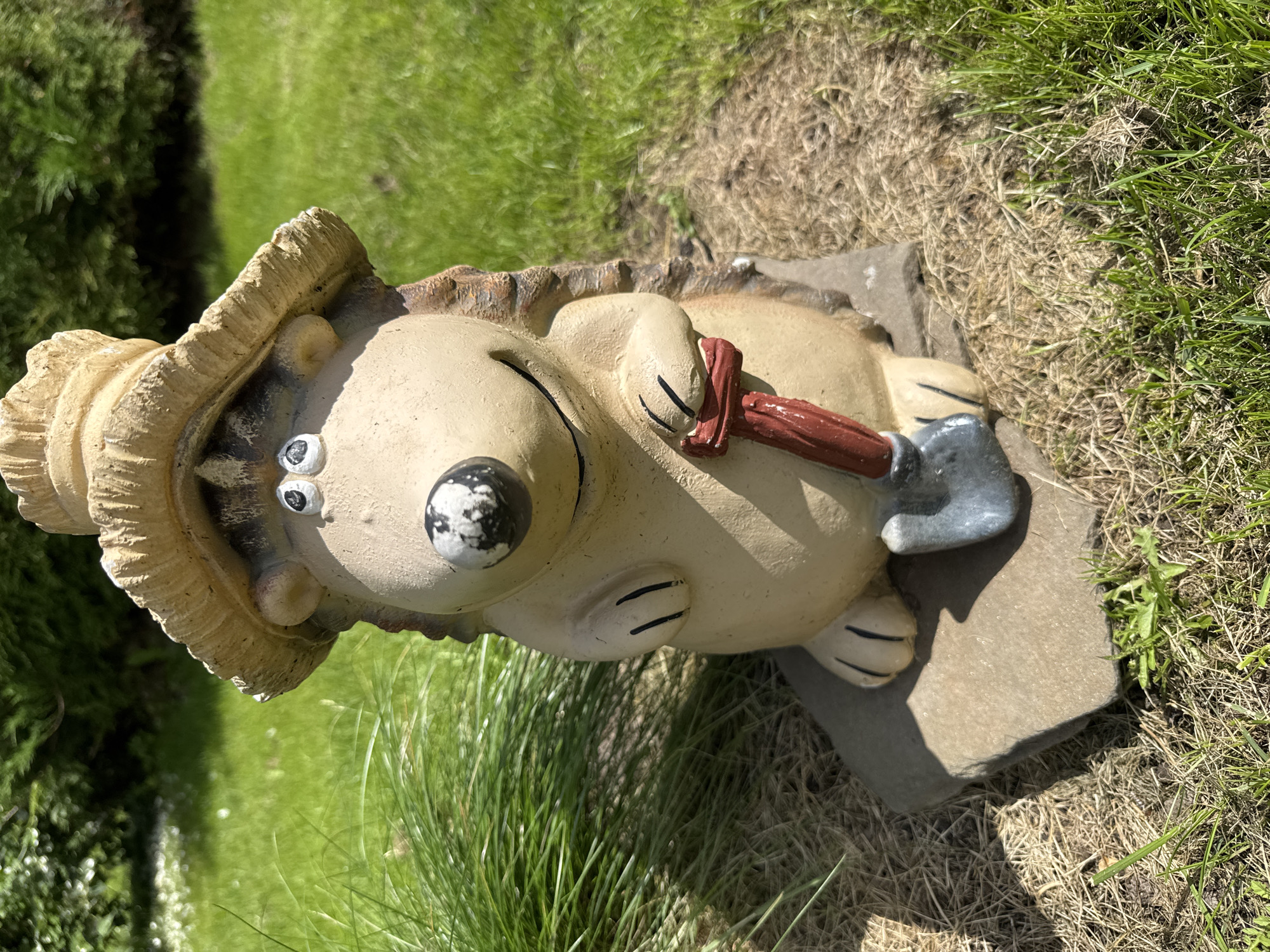 A close-up, eye-level shot features a garden gnome shaped like a hedgehog, standing outdoors on a sunny day in Земледелец-90 Спк, Russia. The gnome is made of a light-colored ceramic material and is wearing a straw hat. It has a round, cream-colored body with black markings on its nose and eyes. Its mouth is curved into a smile, and it has two small, black claws on each hand. The gnome is holding a miniature shovel with a red handle and a gray blade. It is positioned on top of two flat, irregular-shaped stones, which are resting on dry grass and soil. In the background, lush green grass and some out-of-focus foliage are visible, indicating a garden or park setting. The sunlight casts shadows on the gnome and the ground.