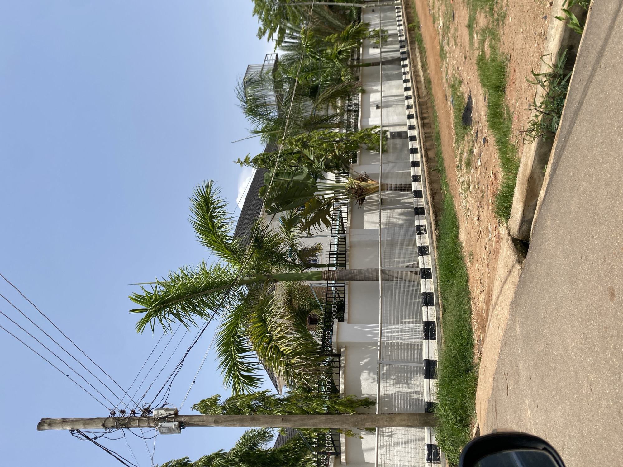 The image depicts a street scene in Gura Top, Nigeria, on a clear, sunny day. A paved road occupies the foreground, with a dirt shoulder and a drainage ditch to the right. Beyond the shoulder, a white, stucco wall encloses a property, topped with a black metal fence featuring ornate circular designs. Lush green trees, including palm trees and other tropical foliage, are visible behind the fence, partially obscuring a modern house with a dark roof. A wooden utility pole with electrical wires and meters stands prominently on the left side of the road. The overall environment suggests a residential area with well-maintained landscaping.