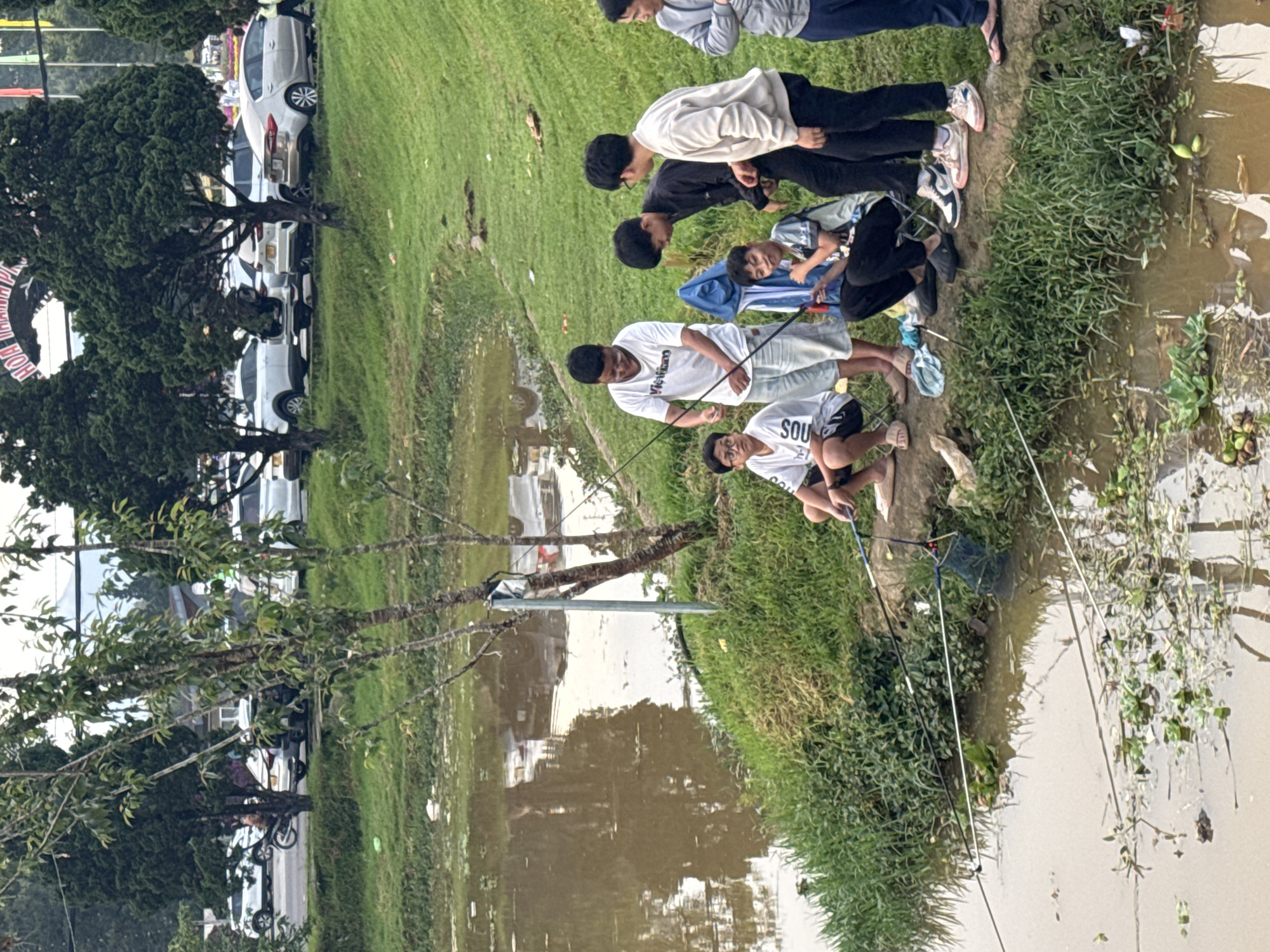A group of at least eight people are fishing along the bank of a body of water; the location is Da Lat, Vietnam. Seven of the individuals appear to be adolescents or young adults, and one is lying down behind them. The people are wearing casual clothing, including t-shirts and pants. They are using fishing rods and nets. The water is murky and brown, and there is vegetation growing along the bank. Behind the bank is a grassy area with trees. In the background, cars are parked in rows along a street. A partial sign and flag can also be seen. The day appears overcast.
