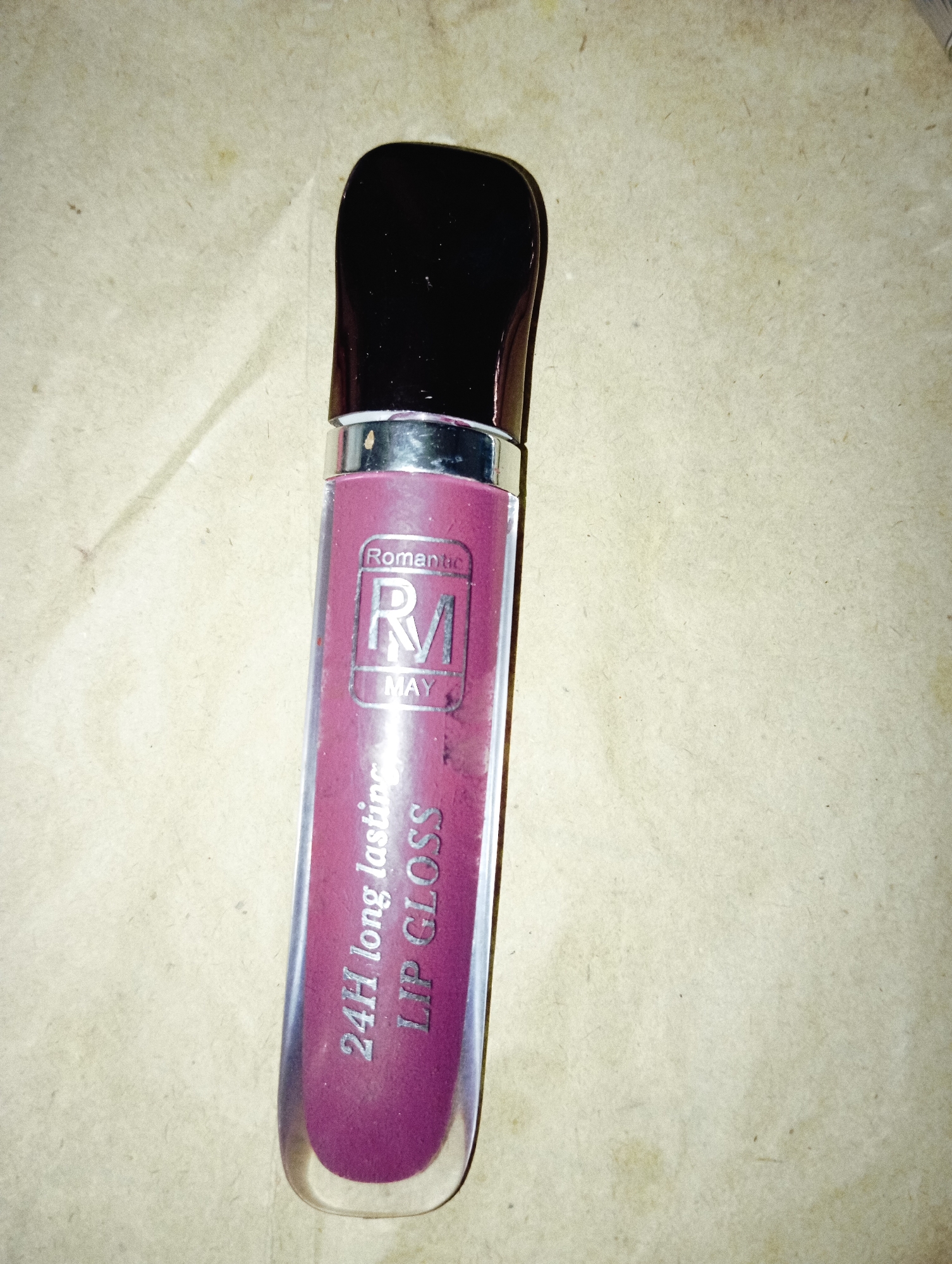 This media file features a close-up, overhead shot of a single tube of lip gloss. The lip gloss tube has a dark, opaque cap and a clear tube that shows the product inside, which appears to be a deep berry or plum shade. The packaging has silver accents and text printed on it.
Visible text on the lip gloss tube includes: "Romantic" written in a script font at the top of a rectangular label. Below that, within the rectangle, is a stylized "RM" logo. Underneath the logo, the word "MAY" is printed. Further down the tube, also printed vertically, are the words "24H long lasting" and "LIP GLOSS".
The background is a textured, light brown surface, possibly paper or cardboard, with some subtle variations in color and a few small specks. The lighting appears to be artificial, casting a slight shadow from the lip gloss tube. No people or specific location cues are visible, and the context suggests a product shot or a casual personal photo. The image focuses solely on the product.