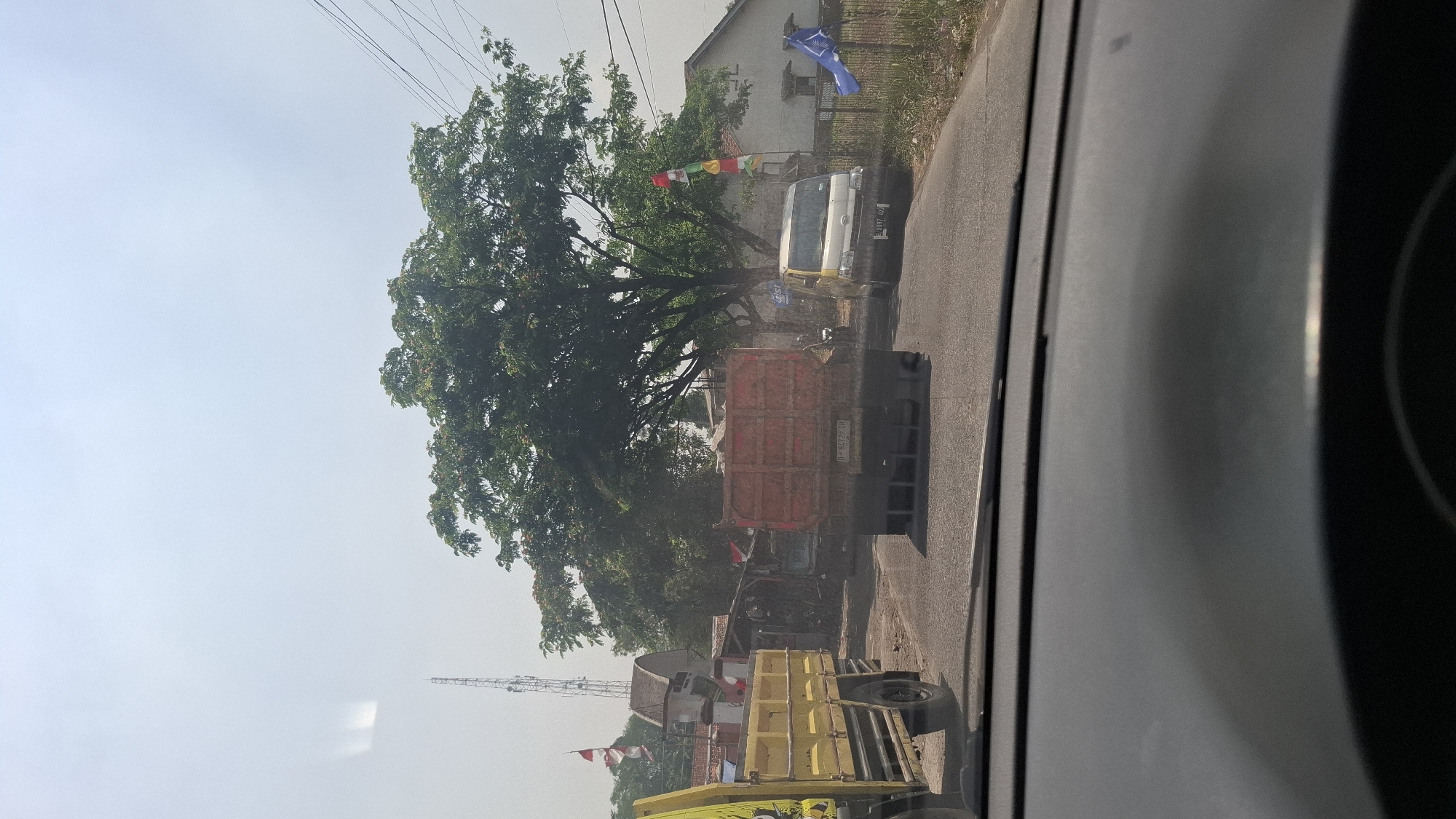 The image captures a street scene in Cibinong Hilir, Indonesia, on a bright, hazy day. The foreground is dominated by the dashboard of a vehicle, implying the photo was taken from inside. In the middle ground, a dirt road is visible, with traffic congestion. Two trucks are prominent: a yellow truck on the left and a red truck in the center. The red truck's license plate reads "D 8472 XR". A white van is parked on the right side of the road. In the background, a large tree with lush green foliage stands tall. A telecommunications tower is visible in the distance, to the left of the tree. Several Indonesian flags are displayed on poles, suggesting a public holiday or event. The environment appears to be semi-rural, with some buildings and vegetation lining the roadside. The sky is pale and washed out, indicating hazy conditions.
