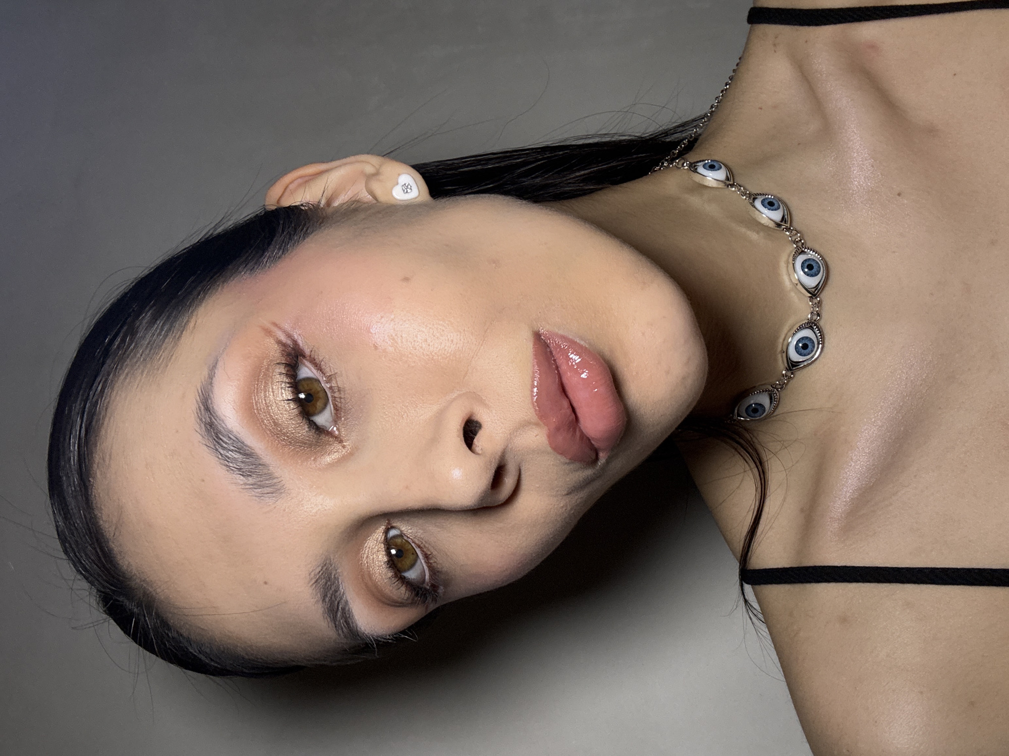 A close-up portrait shows a woman with a light complexion and wet dark hair. Her makeup includes golden eyeshadow, defined eyebrows, and glossy lips. She wears a silver necklace adorned with eye-shaped charms. On her ear, there is a small white heart-shaped sticker or design. The background is a simple, neutral gray. No specific location in Votkinsk, Russia, can be determined from the image.