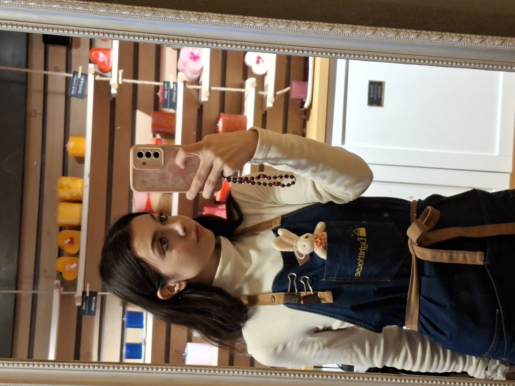 A mirror selfie of a woman wearing a white turtleneck sweater and a dark denim apron with brown suede straps tied around her waist. The apron has a small plush rabbit clipped into the pocket. She is holding a smartphone with a floral-patterned case in her right hand. A string of dark beads wraps around her wrist. The reflection shows a display of vertical wooden slats. Candles of various colors are attached to the slats by small clips. There is a white door visible to the right, with a small black sign mounted on it. The location is a store in an unknown city.