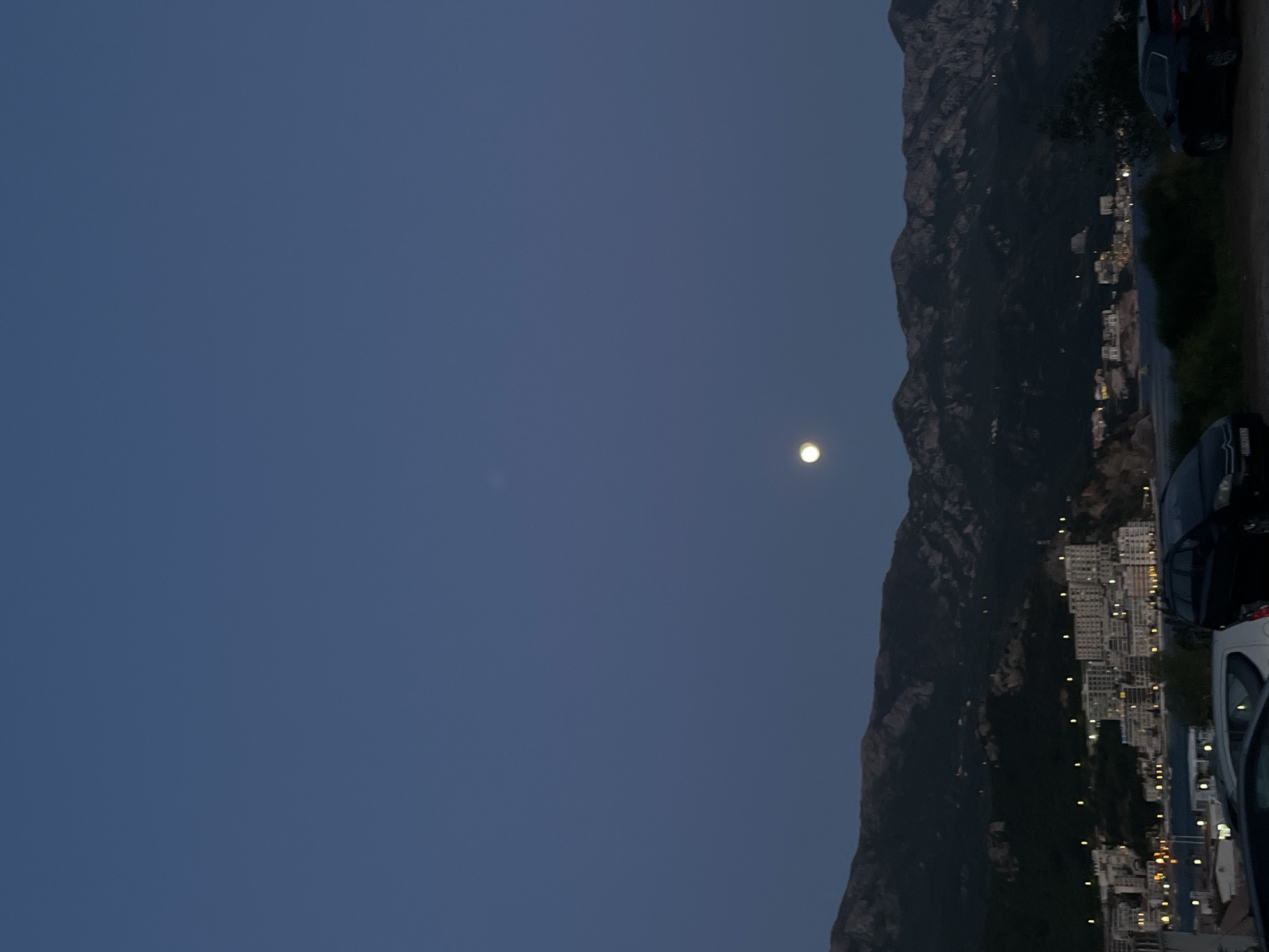The image, taken at dusk or early night, captures a serene coastal scene in what appears to be Boreti, Montenegro. The sky occupies the majority of the frame, displaying a deep, rich blue hue, fading slightly towards the horizon. A prominent, bright moon shines clearly in the upper left quadrant of the sky.
A large, dark mountain range dominates the middle ground, its rugged, forested slopes rising steeply. The mountains are silhouetted against the twilight sky, with some rocky textures visible despite the low light.
At the base of the mountains, a town or settlement stretches along the coastline. Numerous buildings, many of them appearing as multi-story structures, are illuminated with warm, artificial lights, creating a sparkling tapestry against the dark landscape. These lights extend along the shore, with reflections visible on the calm body of water (likely the Adriatic Sea) beyond the town.
In the foreground, parts of several parked cars are visible, indicating a roadside or parking area. A dark vehicle is partially seen on the right, and another on the bottom edge, suggesting the perspective is from a street-level vantage point.
The scene conveys a peaceful atmosphere, highlighting the natural beauty of the mountains and sea alongside the gentle glow of human habitation under a clear, moonlit sky. No people or specific activities are discernible, nor is any text visible.