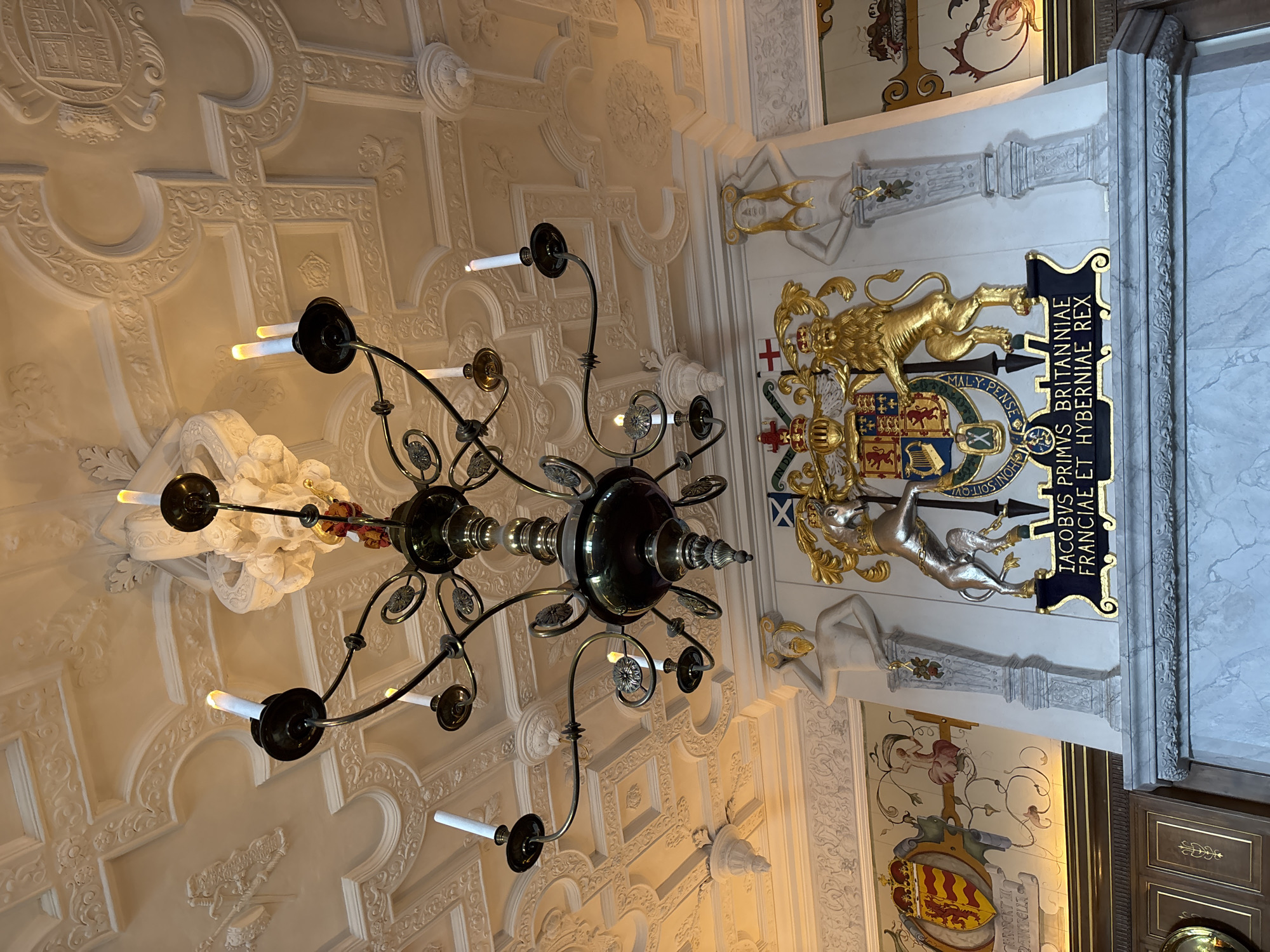 A low-angle shot showcases an ornate ceiling and chandelier in what appears to be a historical building, possibly within the City of Edinburgh, United Kingdom. The ceiling features complex, relief-style decorations and moldings. A black and gold chandelier hangs down, casting a subtle glow from its candle-like lights. Adjacent to the ceiling, a section of the wall is visible with a large coat of arms, flanked by gold statues and artistic paintings, with a frame running vertically in marble.