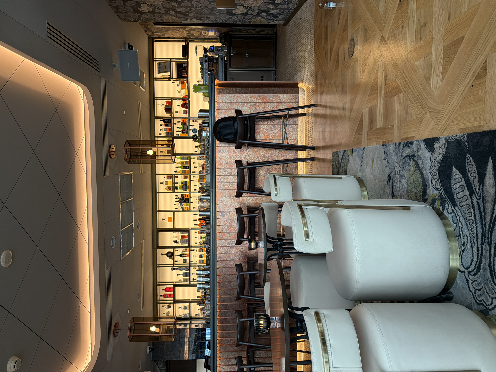 The image depicts an interior shot of a bar. The central focus is a well-stocked bar with illuminated shelving displaying numerous bottles of alcohol. In front of the bar are several bar stools with brown wooden frames. To the right, there's a seating area with white, rounded chairs featuring brass accents. The floor is a patterned wooden design, transitioning into a rug with a floral pattern. The ceiling has recessed lighting and geometric patterns. At the end of the bar, there is a coffee machine. The setting appears upscale and modern. The image does not provide any indication of the location being Sofia, Bulgaria.