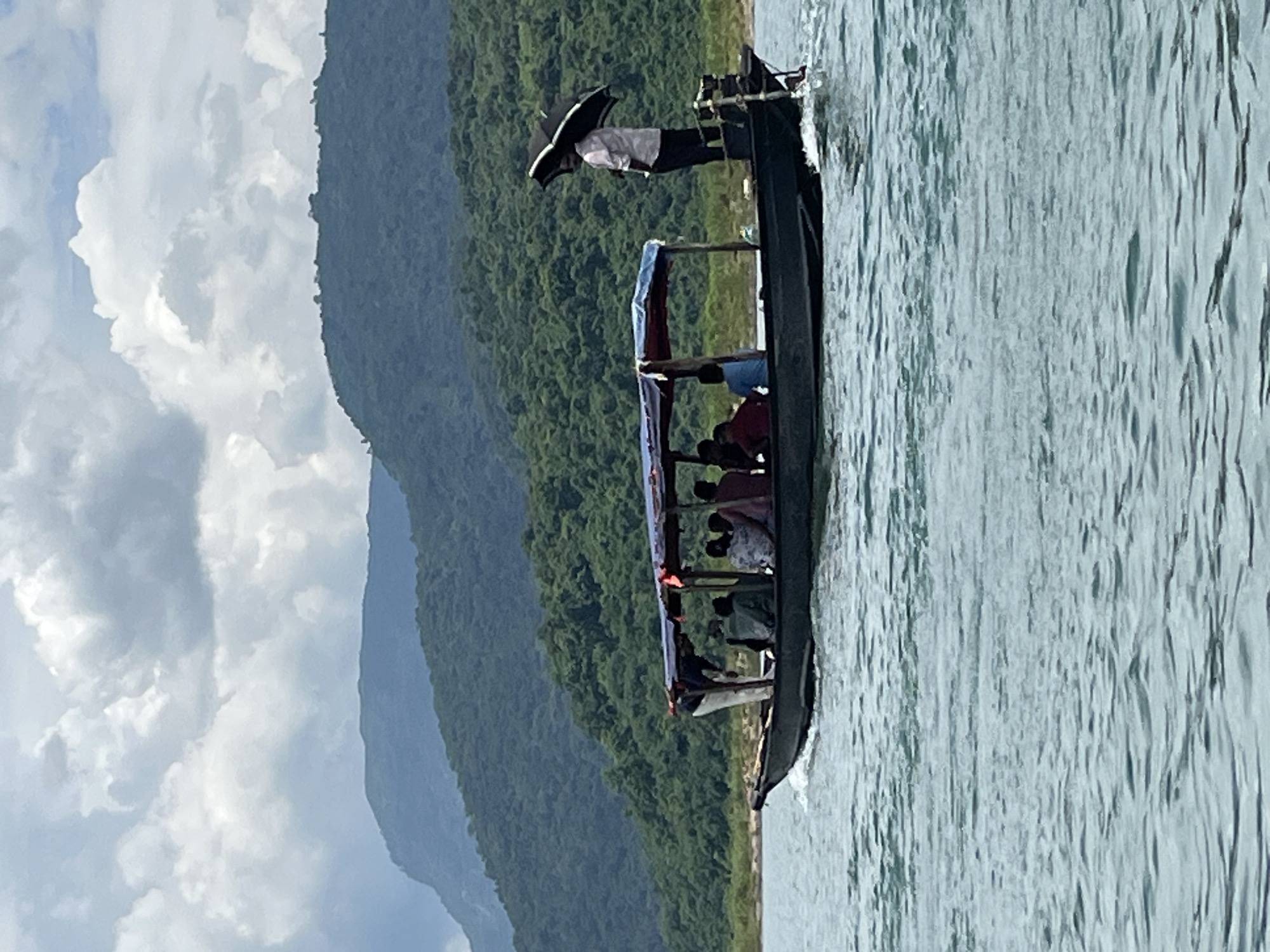 A long, narrow boat filled with several people navigates through calm, rippling water. The boat has a canvas canopy providing shade for the passengers. One person stands at the back of the boat holding a black umbrella. In the background, lush green hills rise up, covered in dense forest, under a cloudy sky. The scene appears to be a scenic view, likely a tourist spot, and given the detail of the hills and vegetation, it is likely in a rural area near Majai, India.