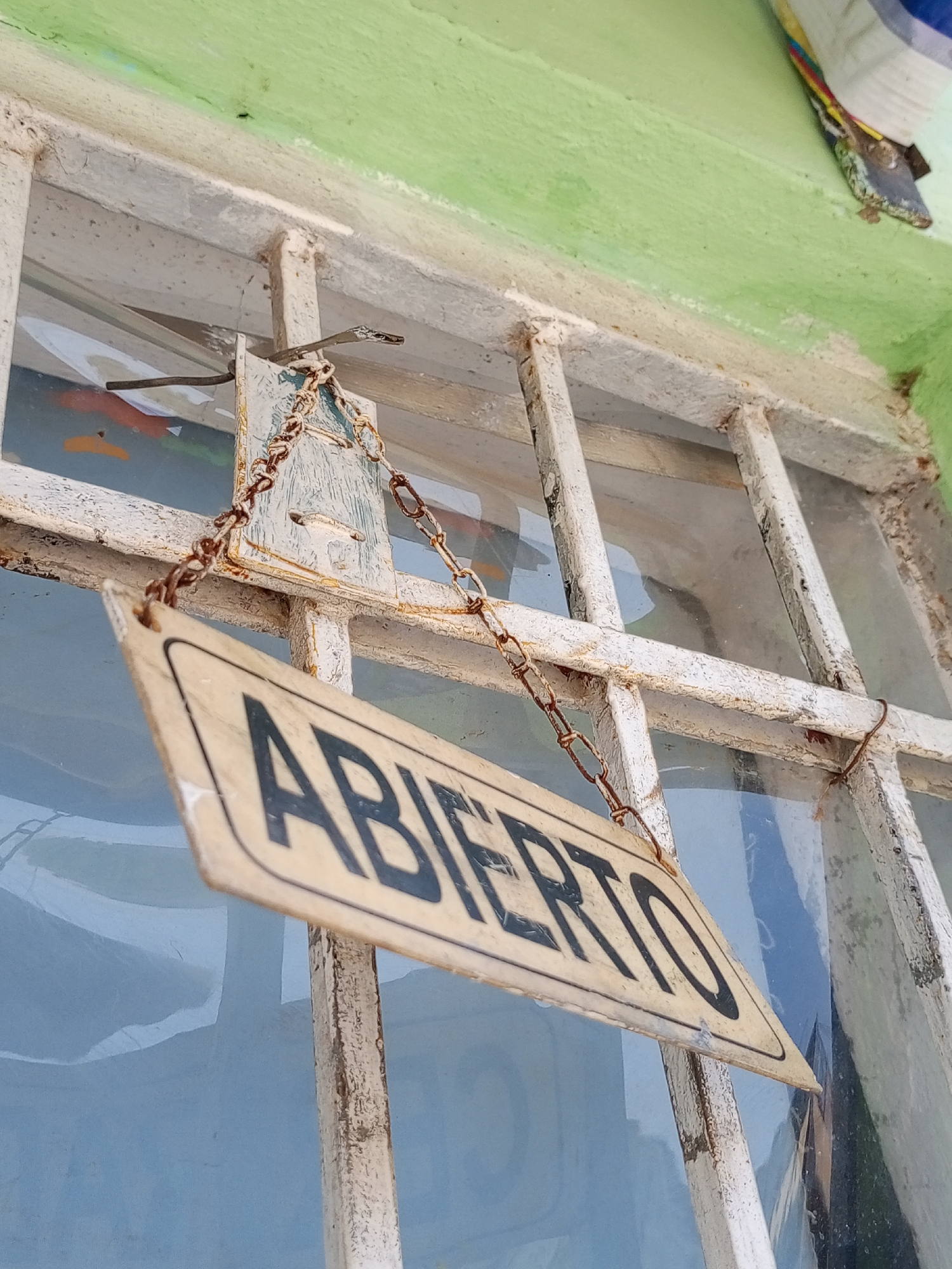 The image depicts an "Abierto" (Open) sign hanging from a weathered, barred window, likely in Porlamar, Venezuela. The sign is rectangular with black lettering on a light-colored background and is suspended by a rusty chain. The window frame is painted white, with visible signs of wear and corrosion. The glass panes are partially obscured by reflections and interior objects. The wall above the window is painted light green, and a lizard is perched on the wall.