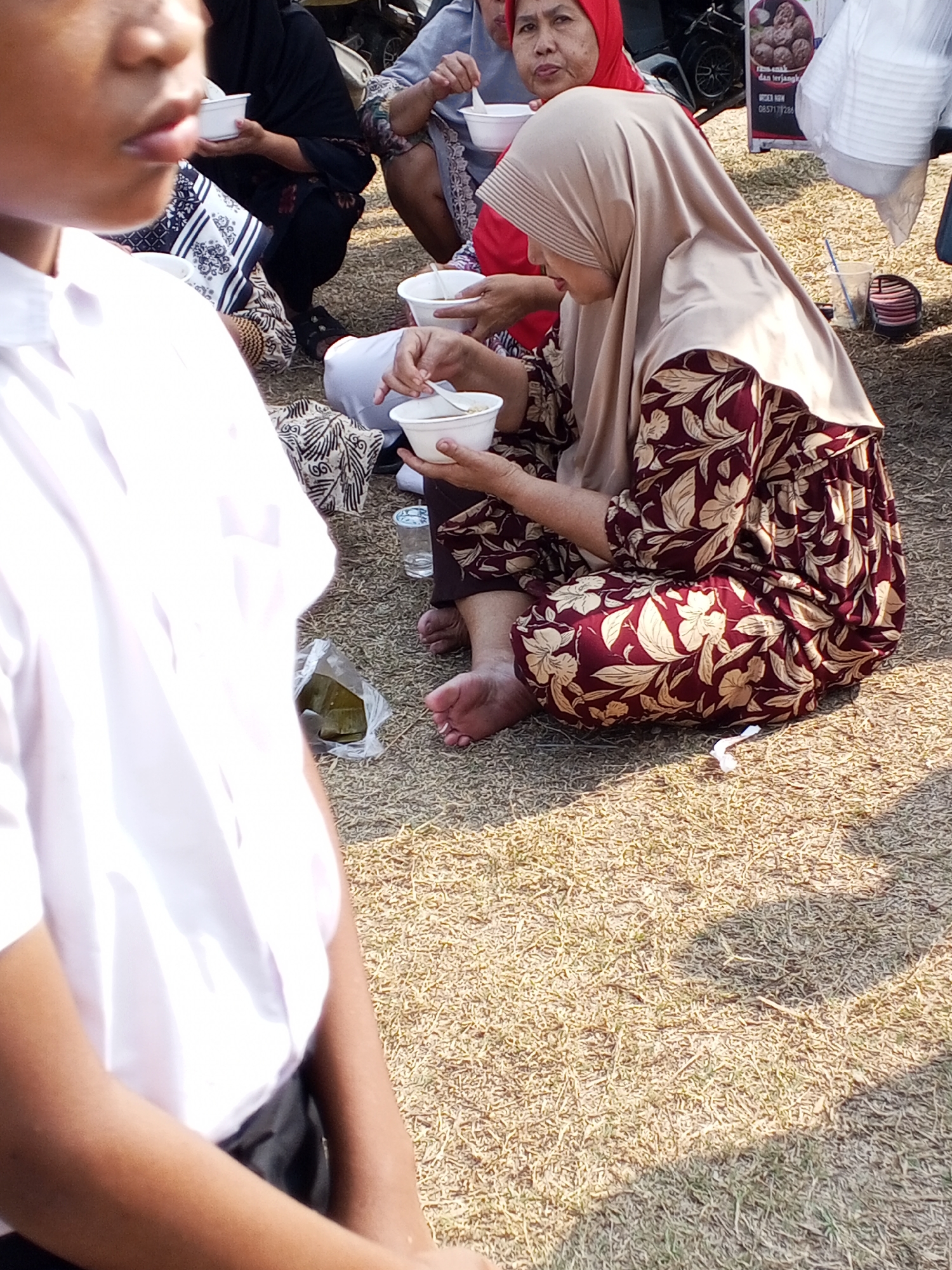 The image depicts an outdoor gathering of people, possibly in Ranjeng, Indonesia, given the context. The foreground is dominated by a young person wearing a white shirt, their face partially visible and looking towards the left. In the middle ground, a woman in a hijab and a patterned maroon and cream dress is seated, eating from a white bowl. Another woman, wearing a red hijab, is also visible in the background, seated and holding a white bowl. The ground is covered with dry grass or straw, suggesting an outdoor event. Several white bowls, a plastic bag containing food, and a disposable cup with a straw are scattered on the ground, indicating a meal or picnic. The lighting suggests it is daytime, with shadows cast by unseen objects.