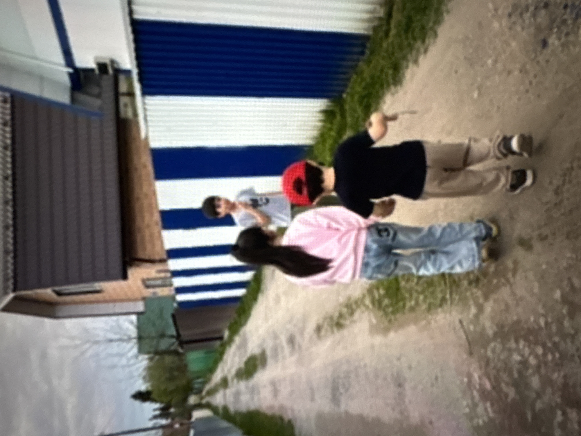 The image shows three individuals standing on a pathway adjacent to a fence in Ембаево, Russia. The pathway is lined with grass. The fence consists of alternating blue and white horizontal stripes. A building with a brown roof is visible in the background. The person closest to the camera is wearing a black shirt and tan pants, and appears to be holding a stick. The middle person wears a red baseball cap and a pink long-sleeve shirt, and jeans. The person farthest from the camera wears a gray t-shirt.